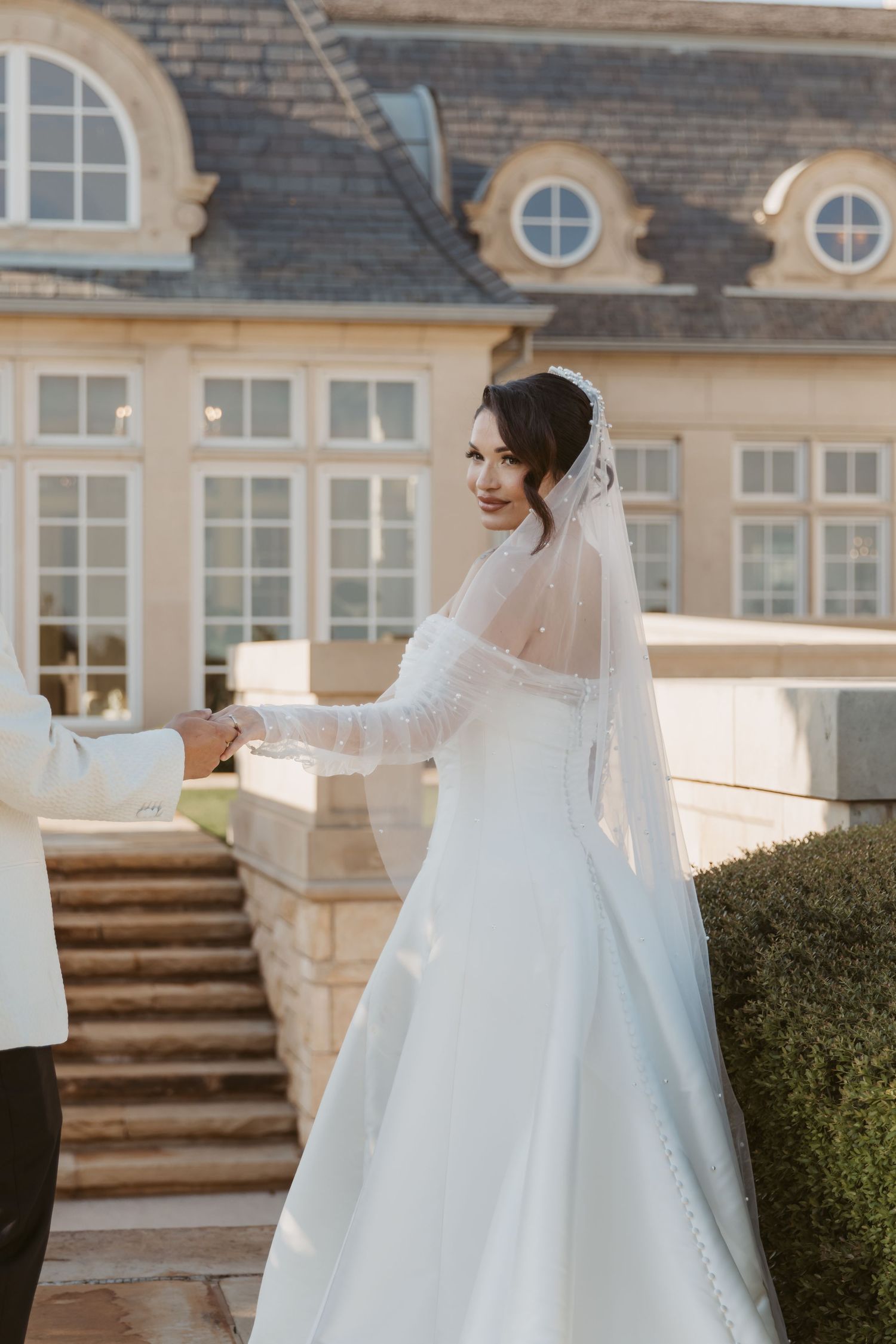 Elegant bride in flowing white gown poses outside a grand mansion with ornate architectural details.
