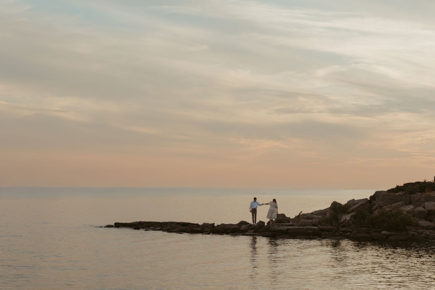 A serene coastal lighthouse perched on rocky cliffs at sunset with soft orange clouds reflecting in the ocean.
