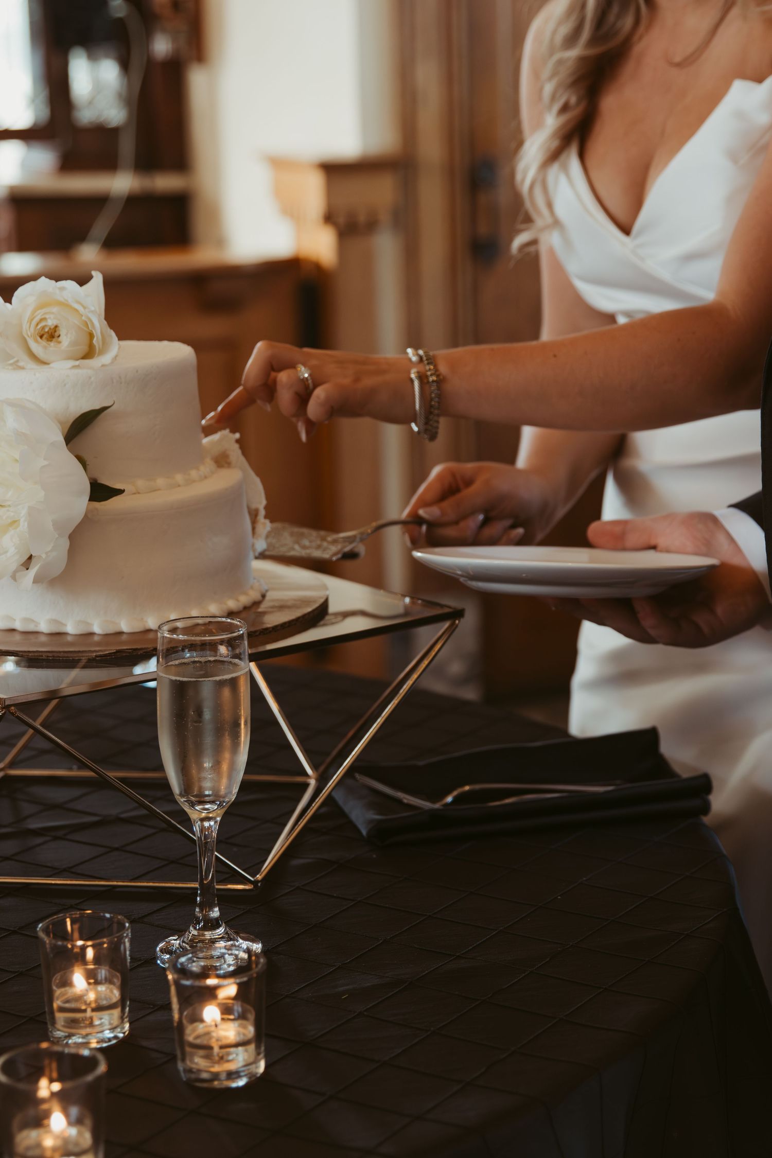 Someone in a white dress cuts into an elegant white wedding cake adorned with roses while champagne glasses sparkle nearby.