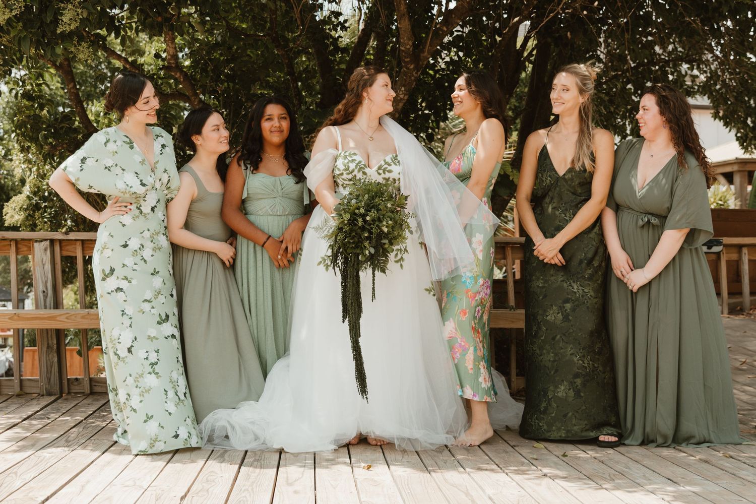 A bridal party in mix-matched green dresses poses together on a wooden deck.