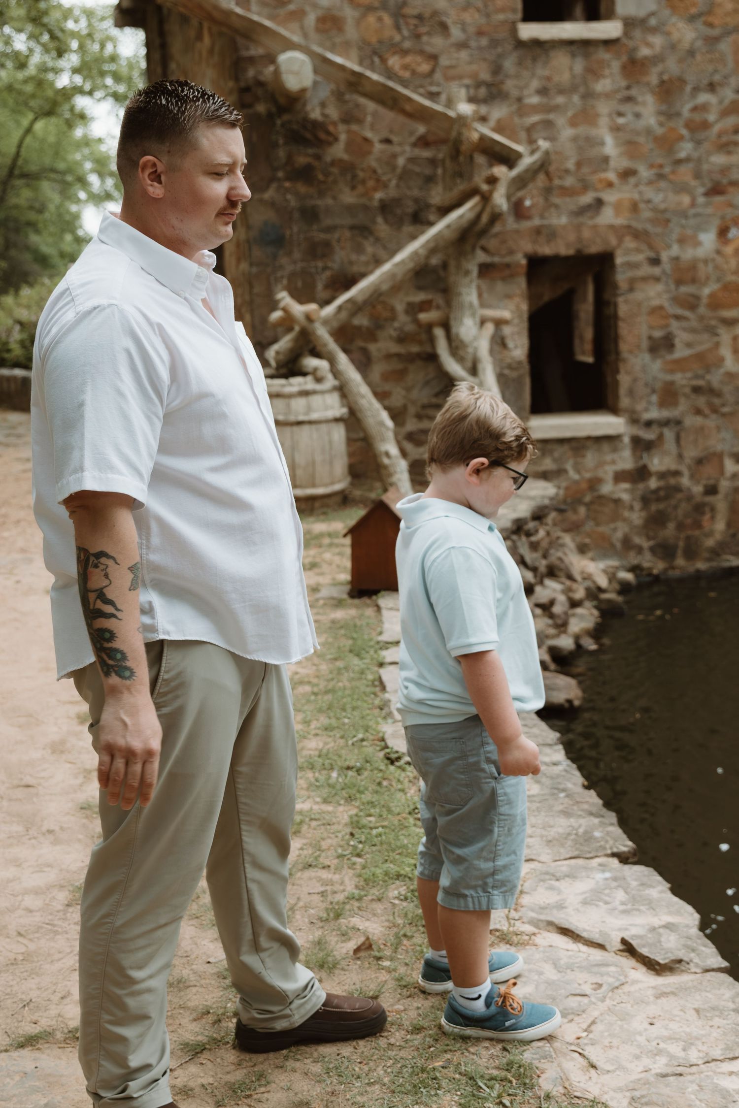 Two people stand next to a historic stone mill building beside a small pond on a sunny afternoon.