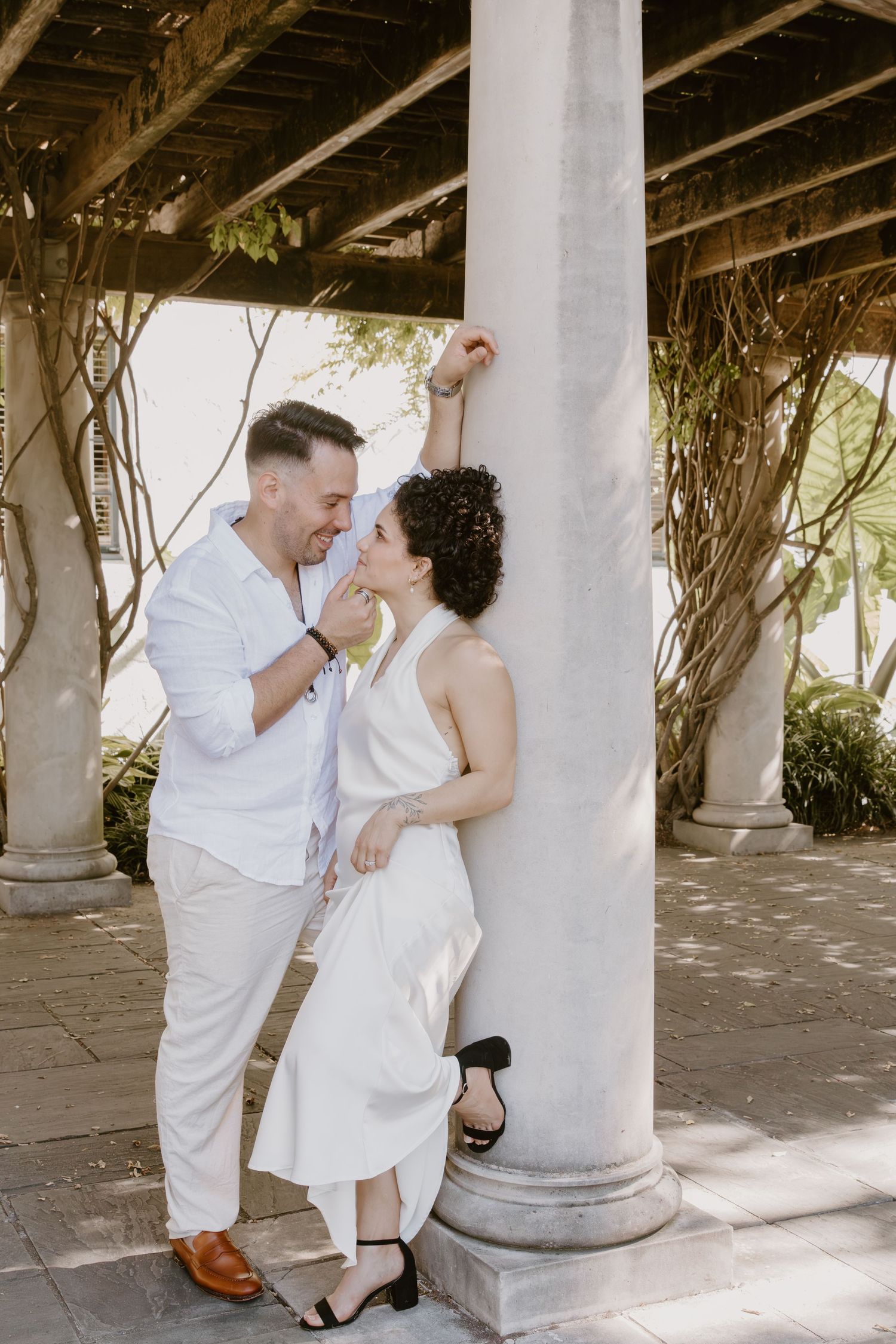 Romantic couple in white attire sharing intimate moments around a classical column in a sun-drenched courtyard.