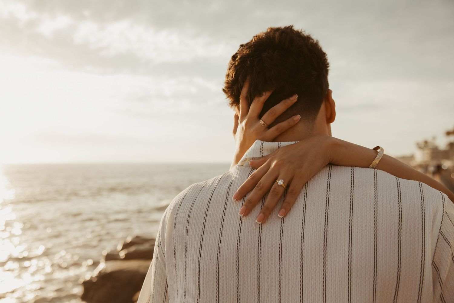 Couple embracing at sunset by the ocean, woman's hand with engagement ring resting on man's shoulder.