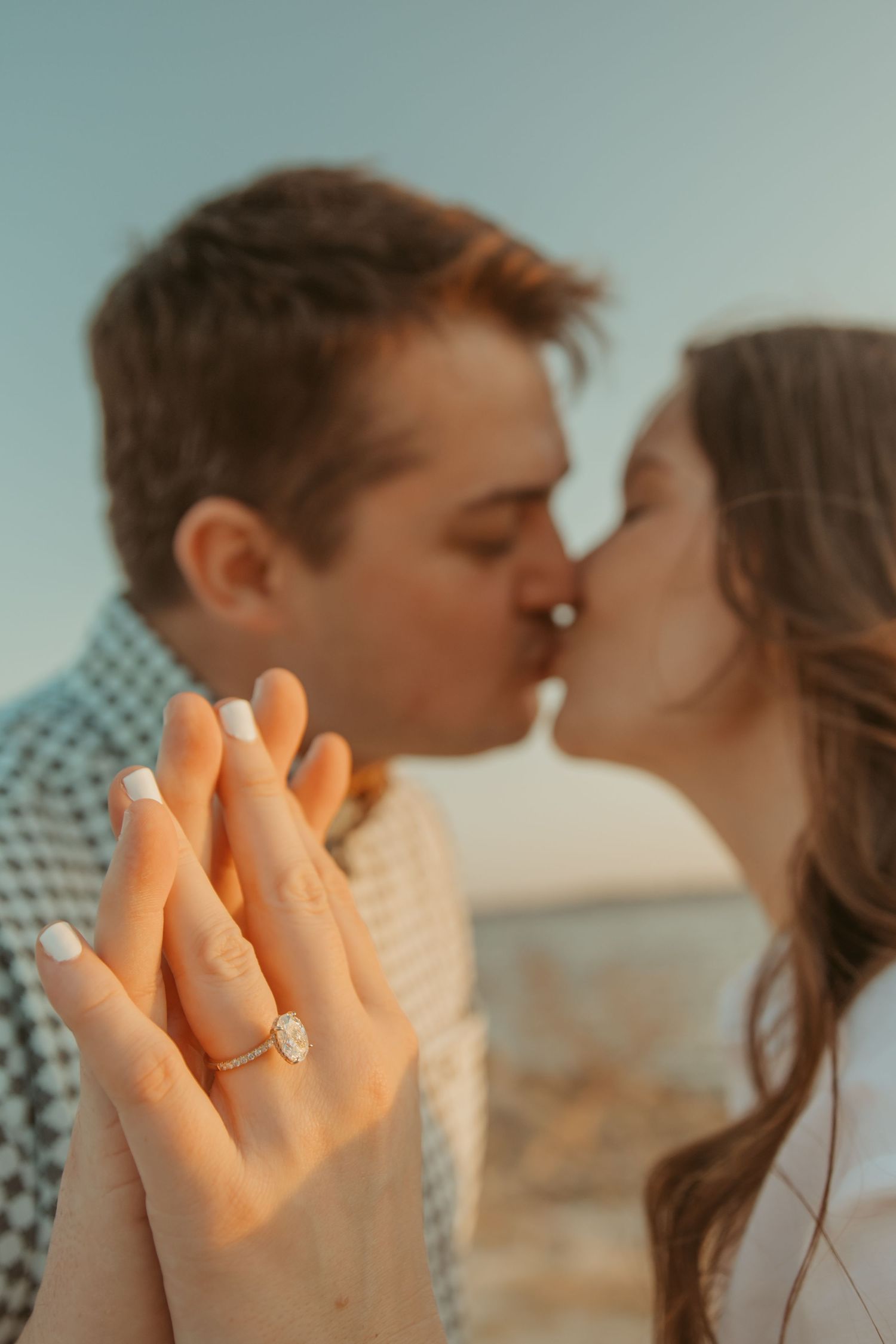 Couple kissing at sunset while showing off a sparkling oval engagement ring in the foreground.