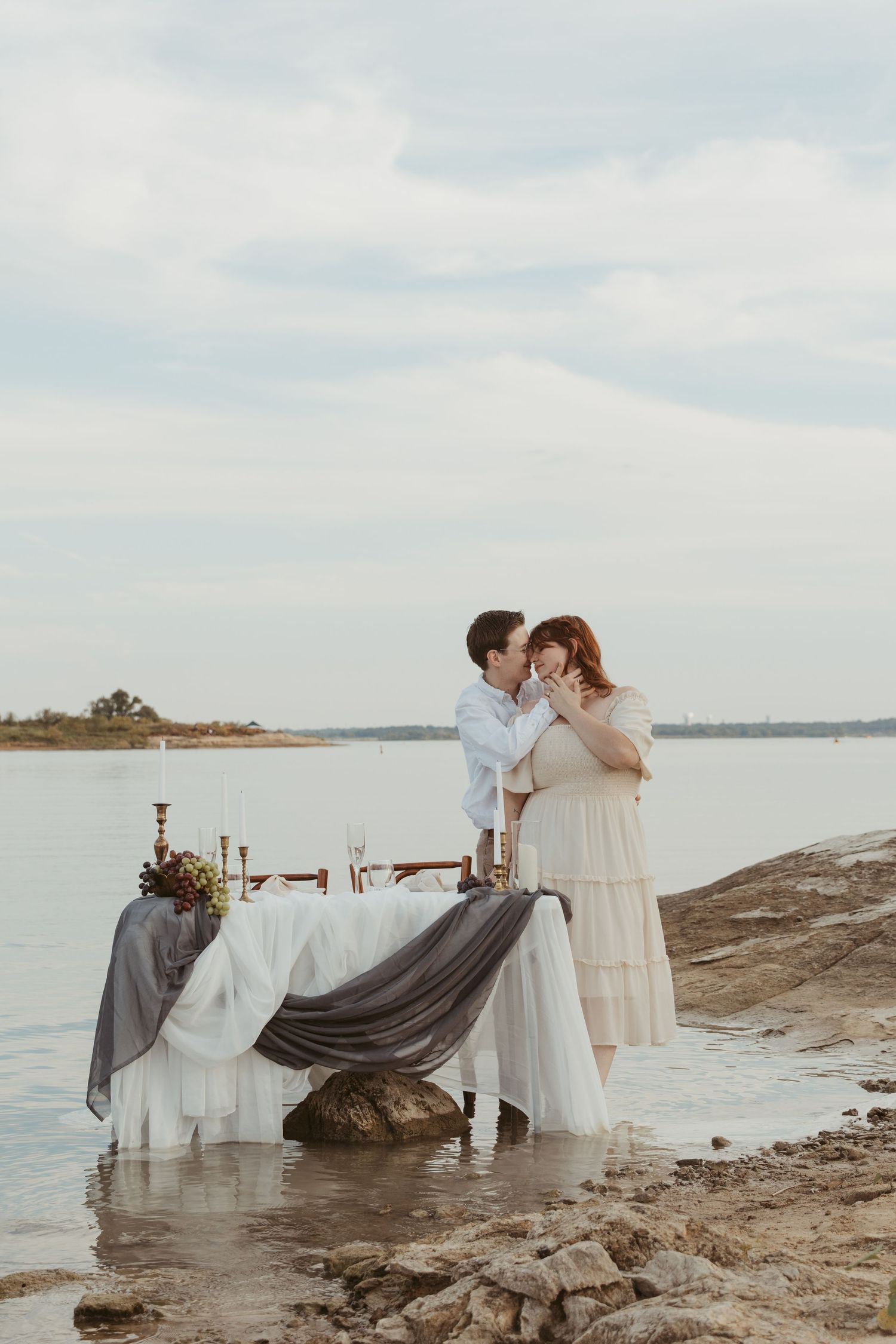 An intimate waterfront scene with a decorated table and couple by the shore at sunset.