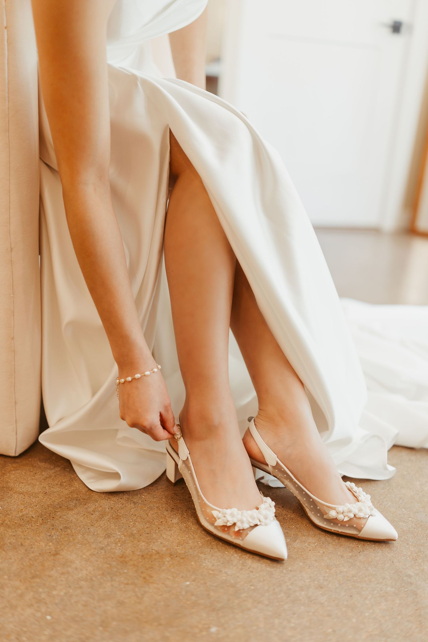 Bride adjusting elegant ivory slingback heels with floral details, wearing a white satin gown and delicate ankle bracelet.