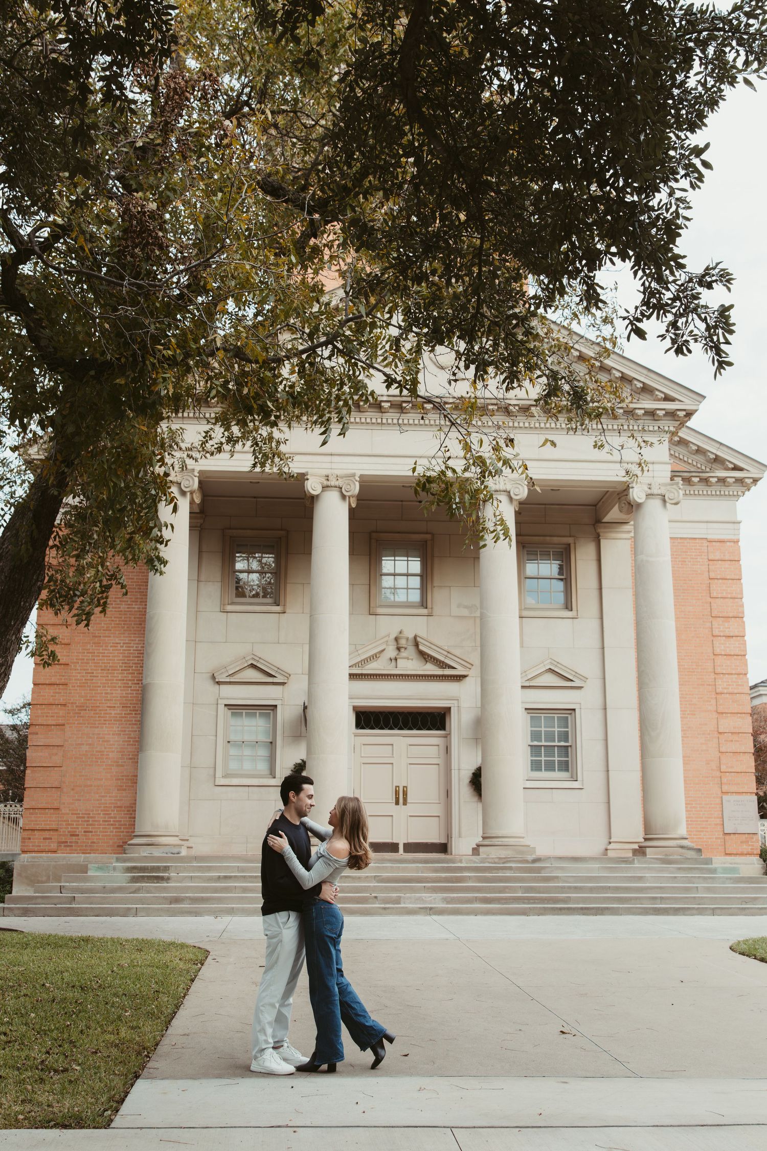 A couple dances playfully in front of a grand neoclassical building with white columns and pink walls. engagement photos Fort Worth