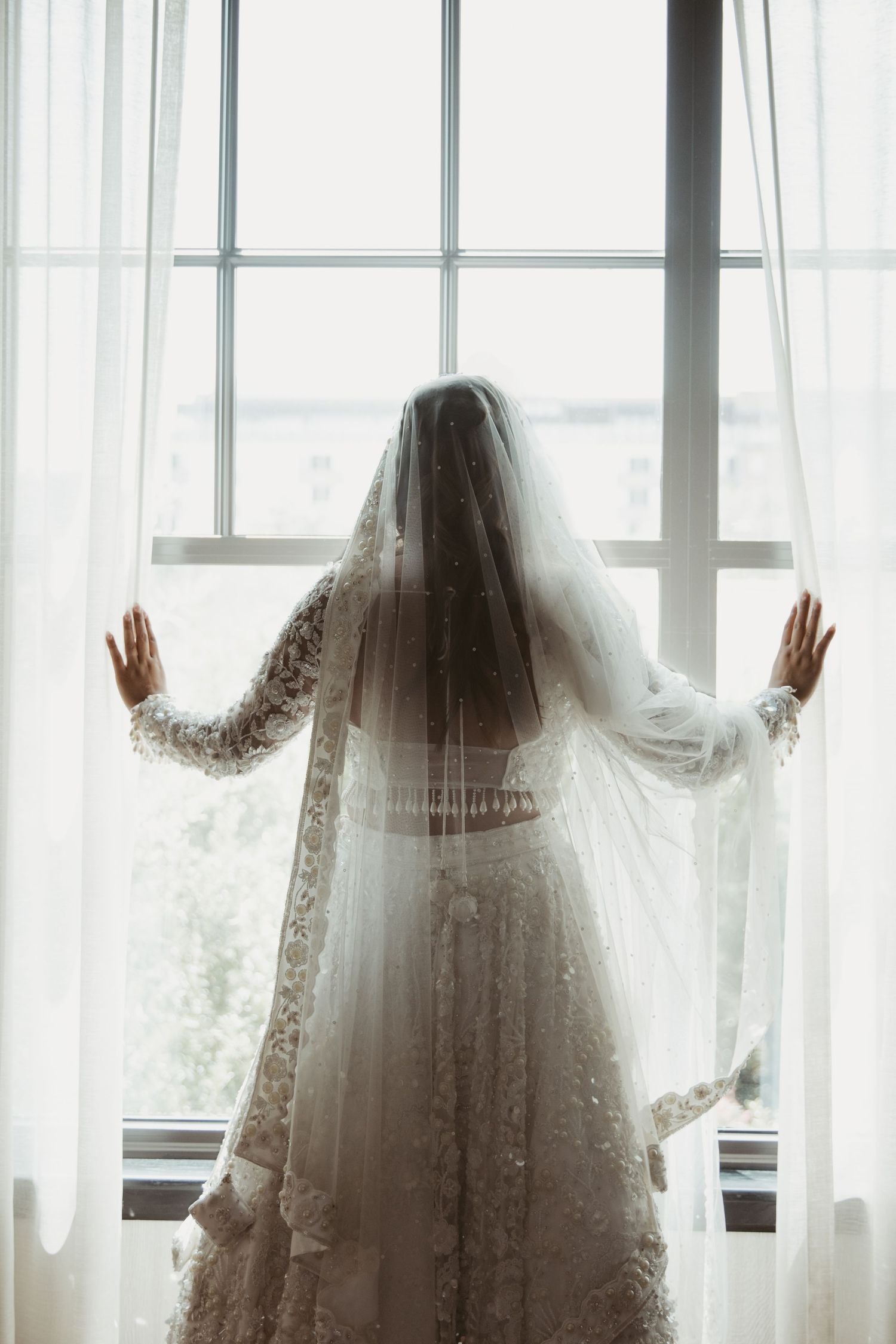 A bride in a lace wedding dress and veil stands silhouetted against a bright window with sheer curtains.