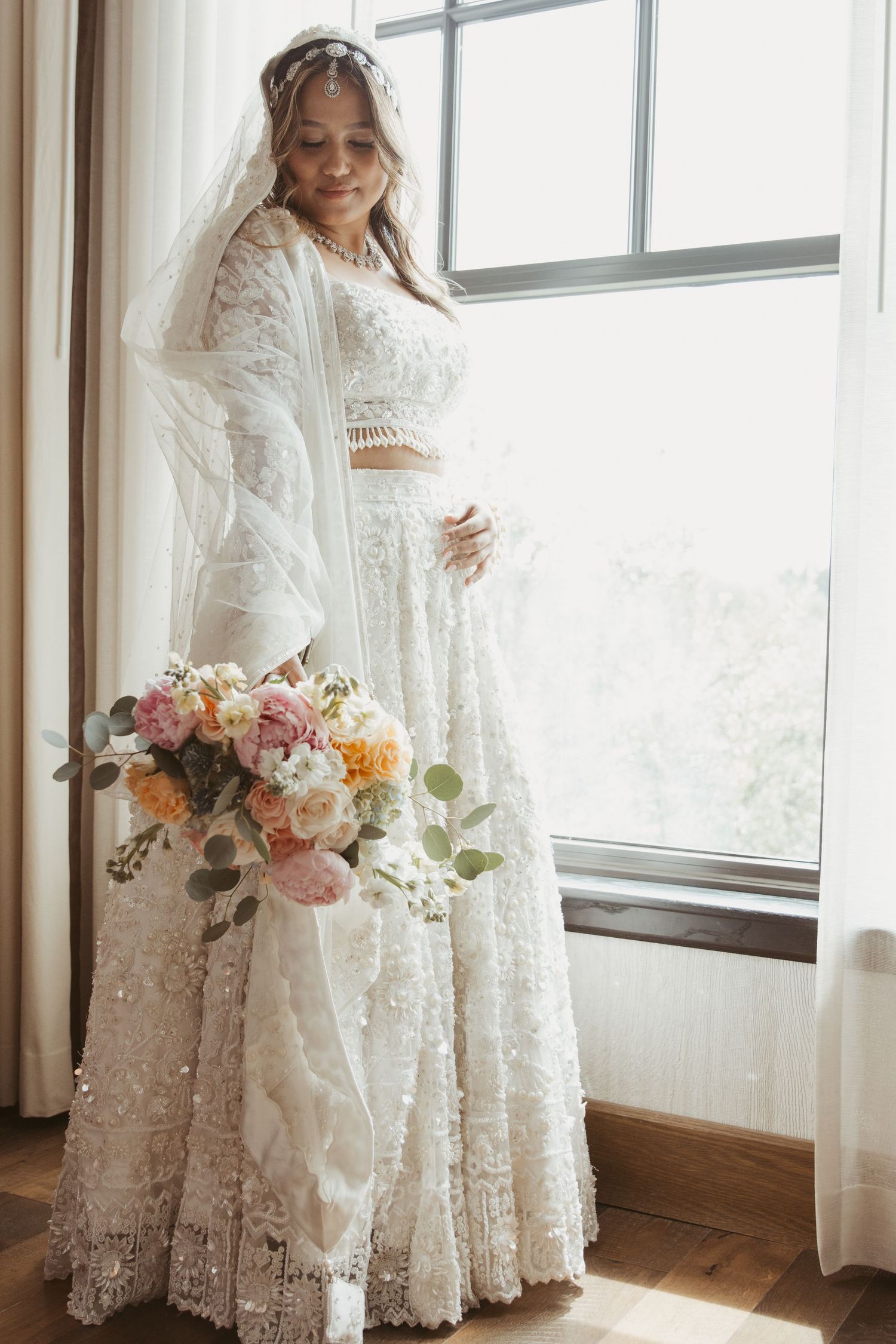 A bride in a two-piece lace wedding dress poses by a window with a colorful bouquet.