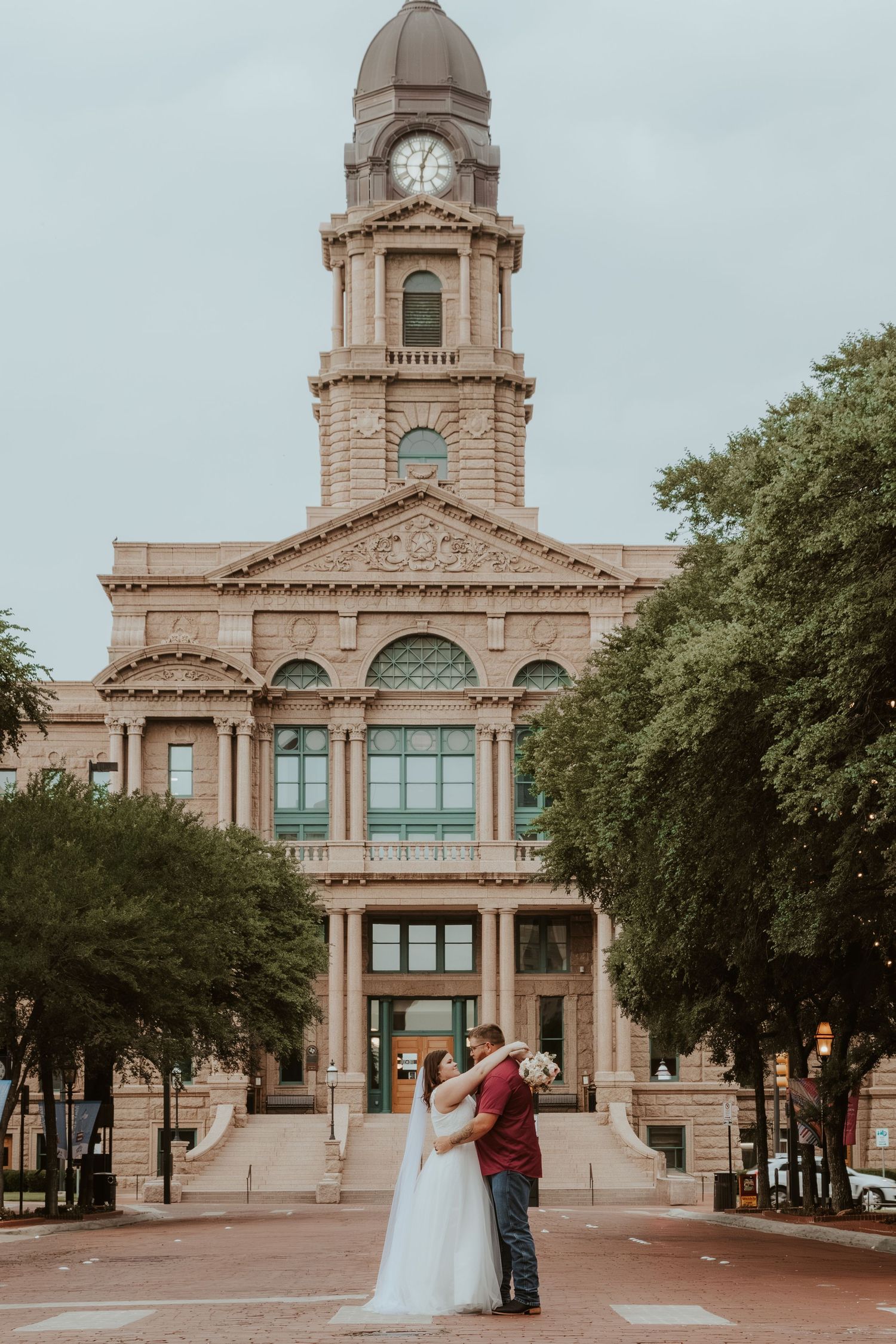 A historic courthouse building with a clock tower serves as the backdrop for a romantic couple in Fort Worth, Texas.