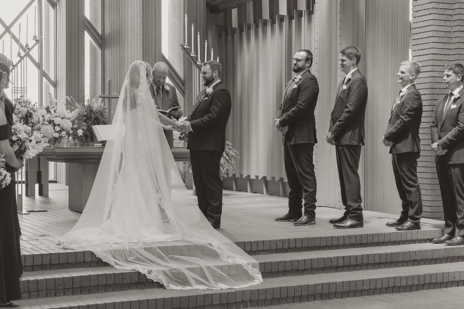 Black and white wedding party lineup shows groomsmen and bridesmaids standing with the newlyweds on a church porch.