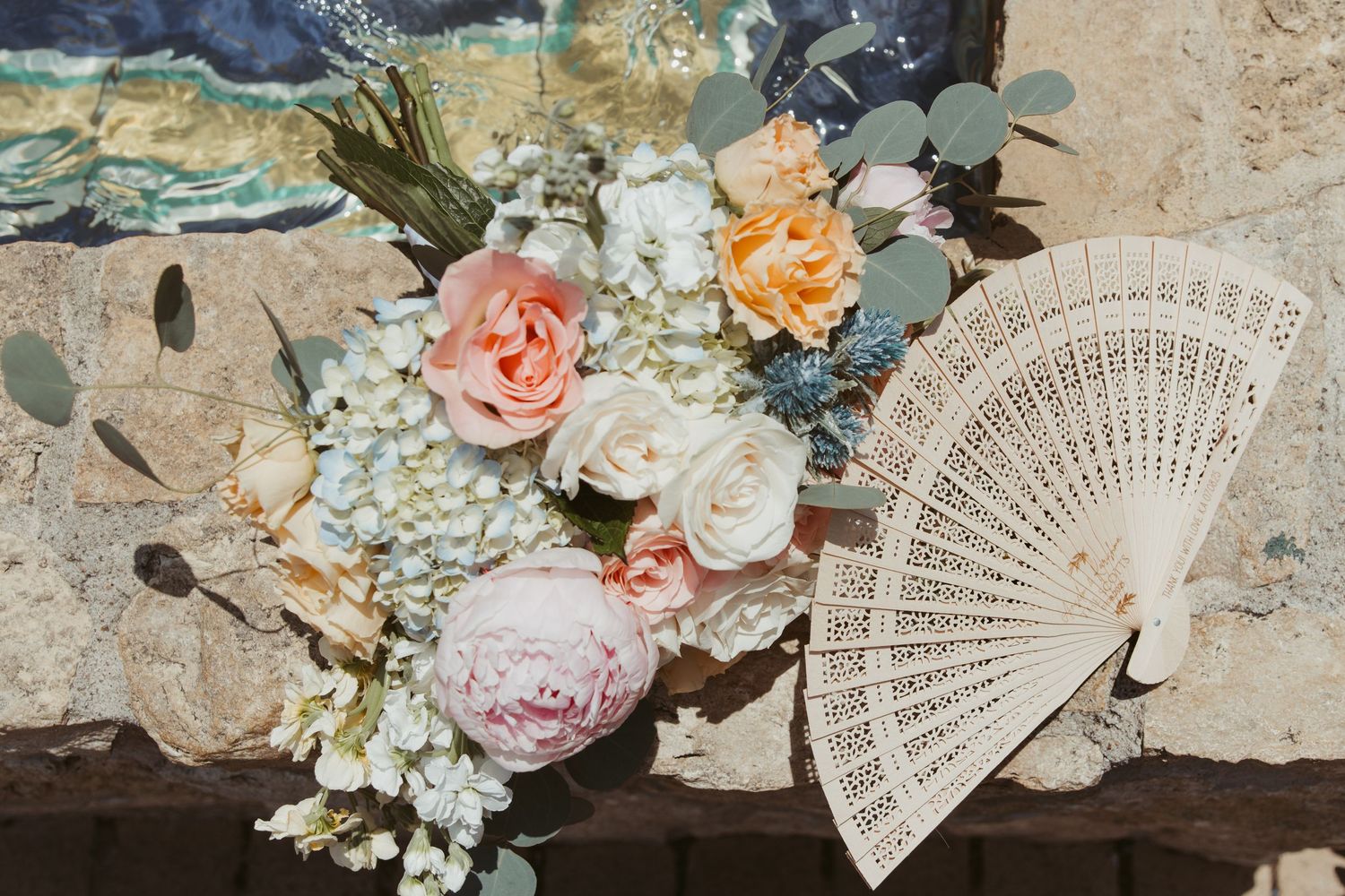A delicate ivory lace fan displayed beside a romantic bouquet of peach, pink and white roses with eucalyptus leaves.