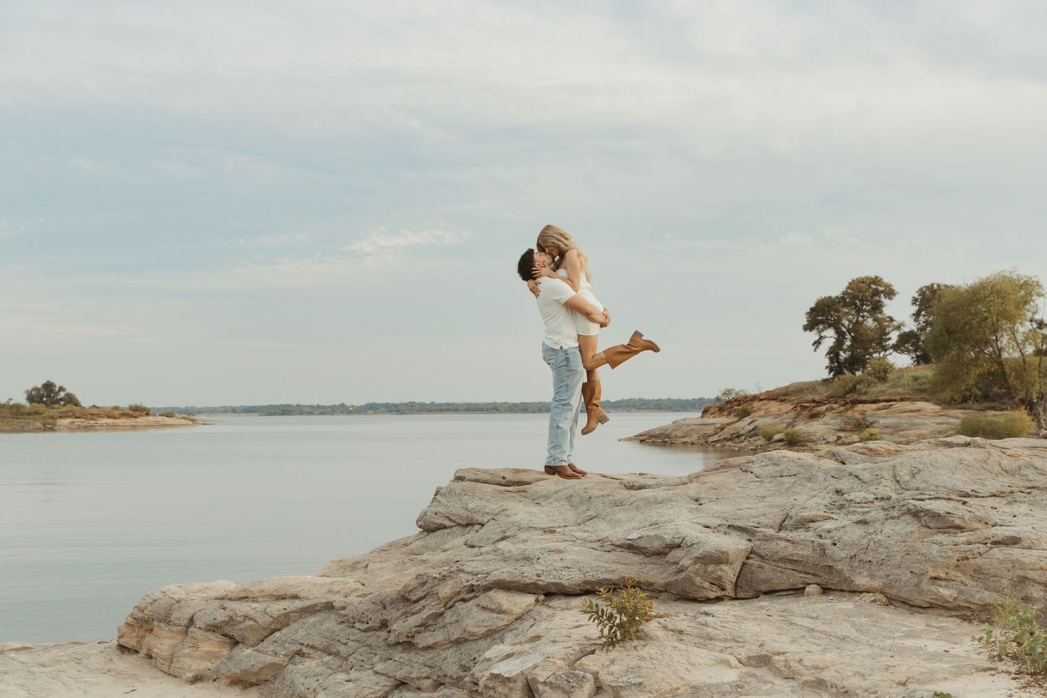 A joyful figure leaps on rocky coastal outcrop overlooking calm waters on a hazy summer day.