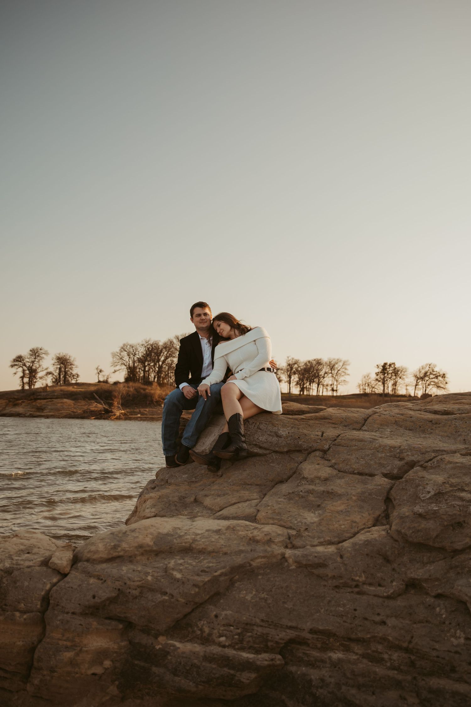 Couple sitting together on rocky lakeside outcrop at sunset, woman leaning on mans shoulder, trees in background.