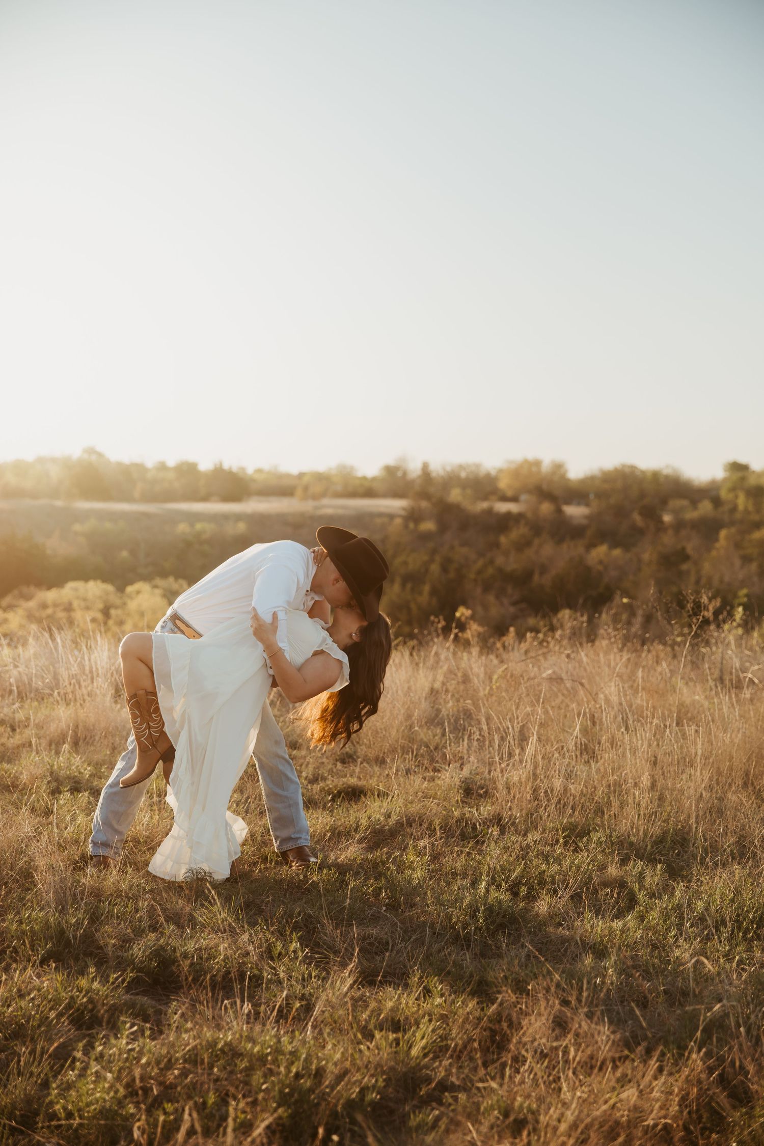 Couple sharing a romantic dip kiss in a golden sunlit field during an outdoor wedding photoshoot.
