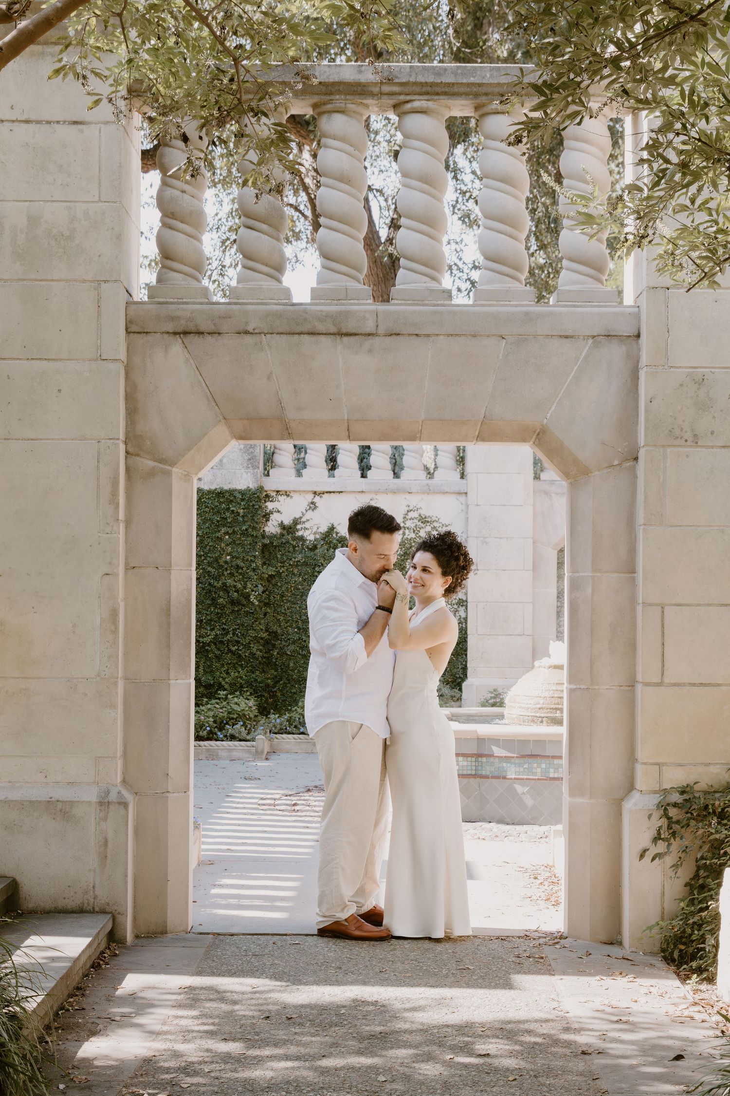A wedding couple shares romantic moments under an elegant stone archway with white balusters.