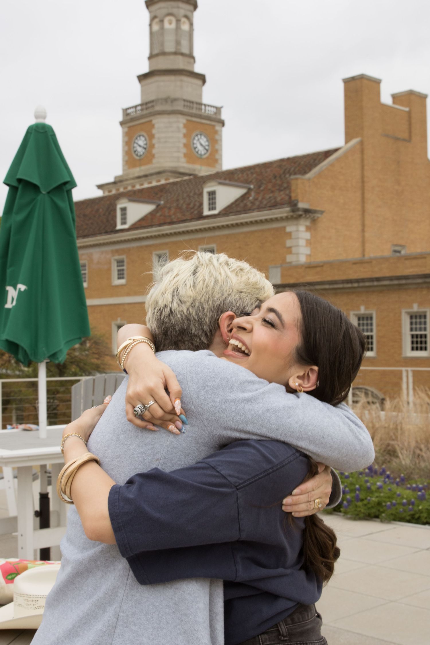 Two people embrace in a warm hug in front of a historic brick building with a clock tower.