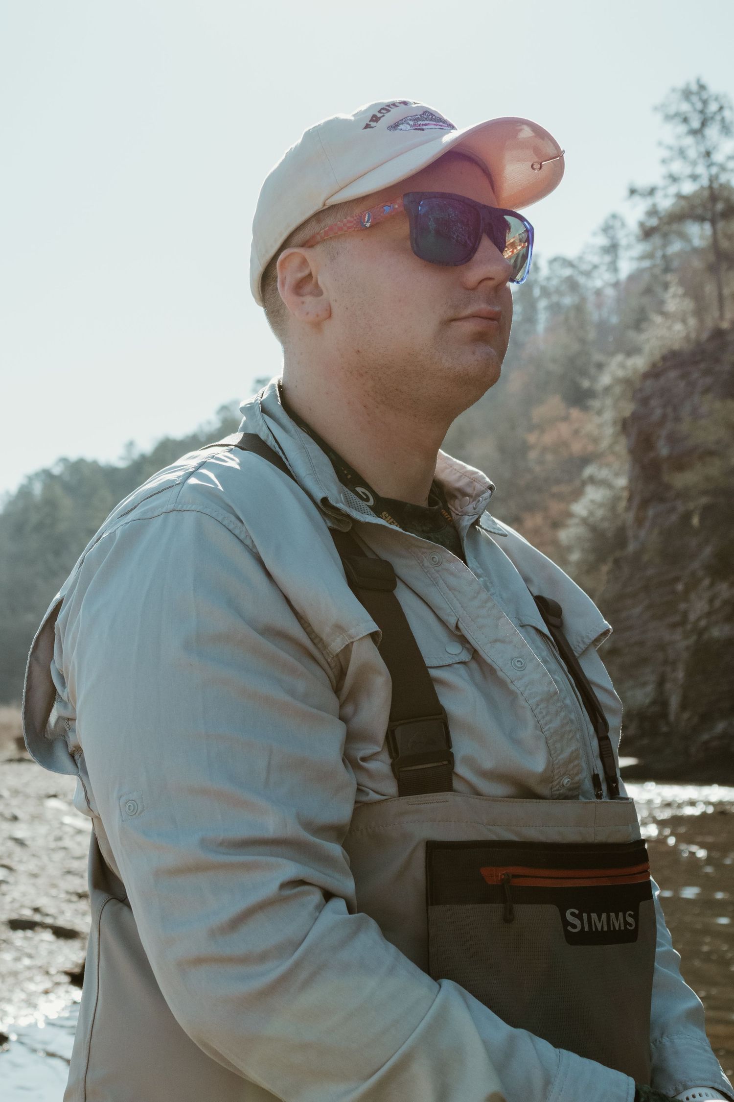 A fly fisher wearing waders and sunglasses stands by a riverbank.