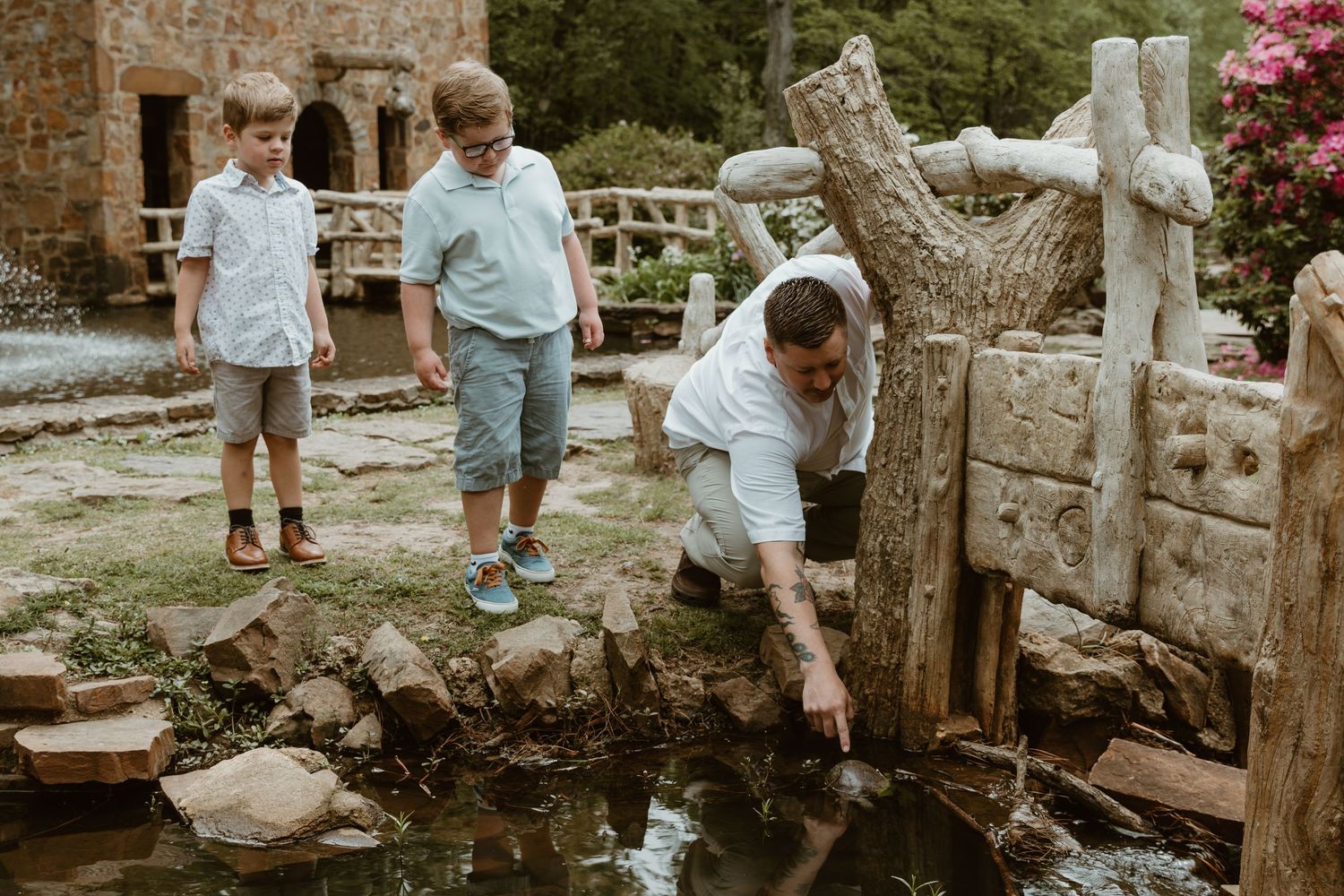 Three young children explore a peaceful garden pond surrounded by stone landscaping and flowering plants.