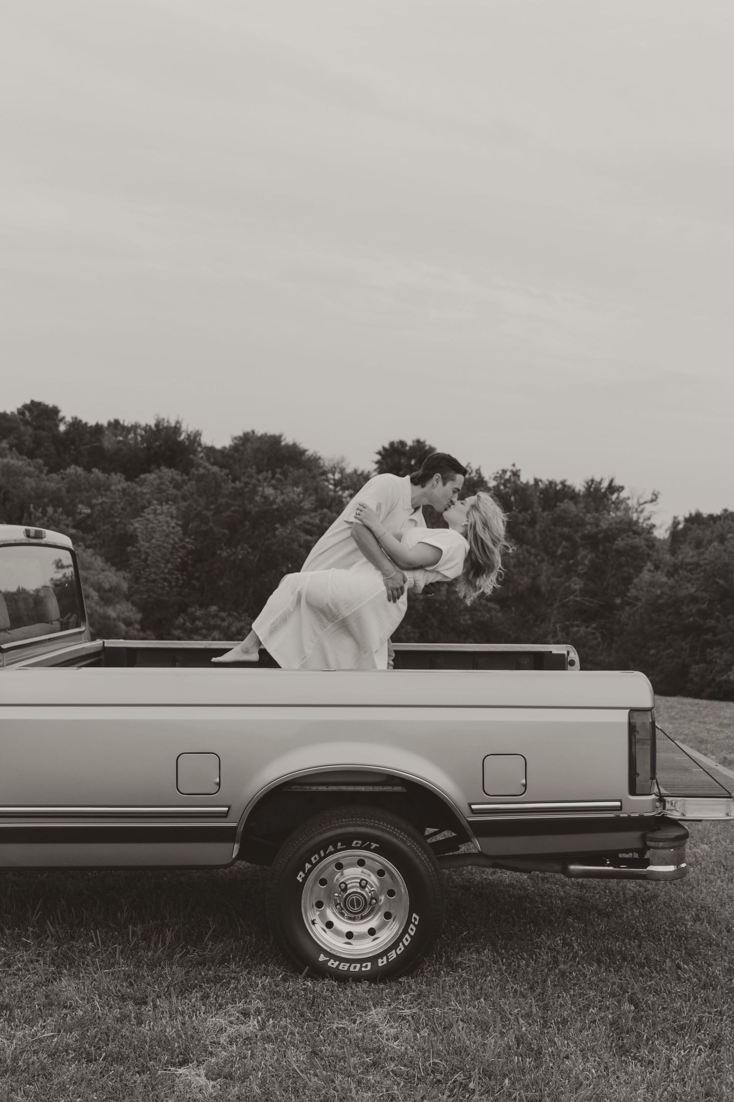 A couple embraces romantically in black and white on the bed of a vintage pickup truck in a countryside setting.