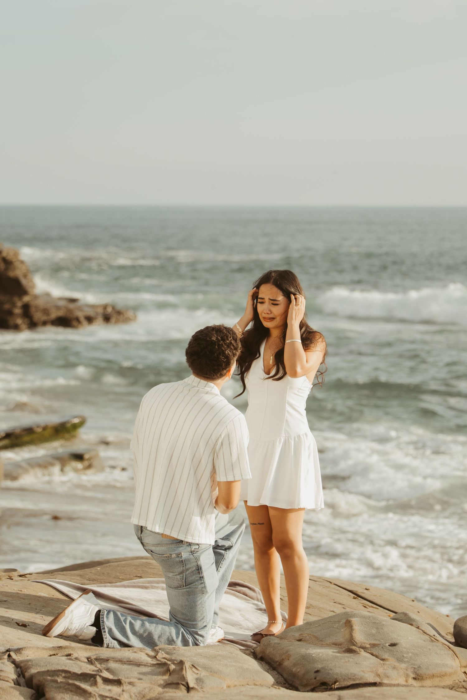 Man kneeling on rocky beach proposing to woman in white dress with ocean waves in background.