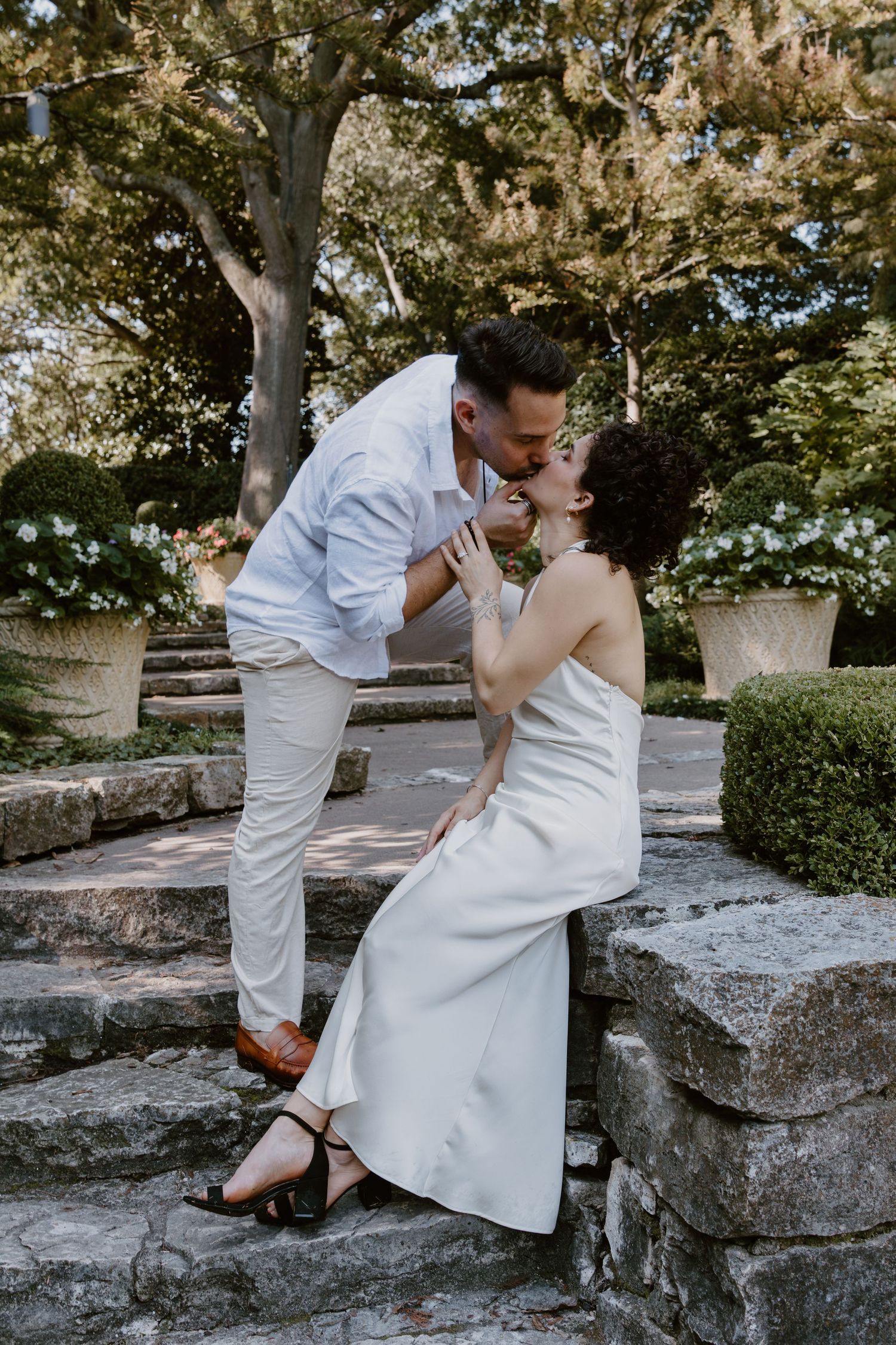 A romantic couple in white outfits shares intimate moments on stone steps in a garden setting.