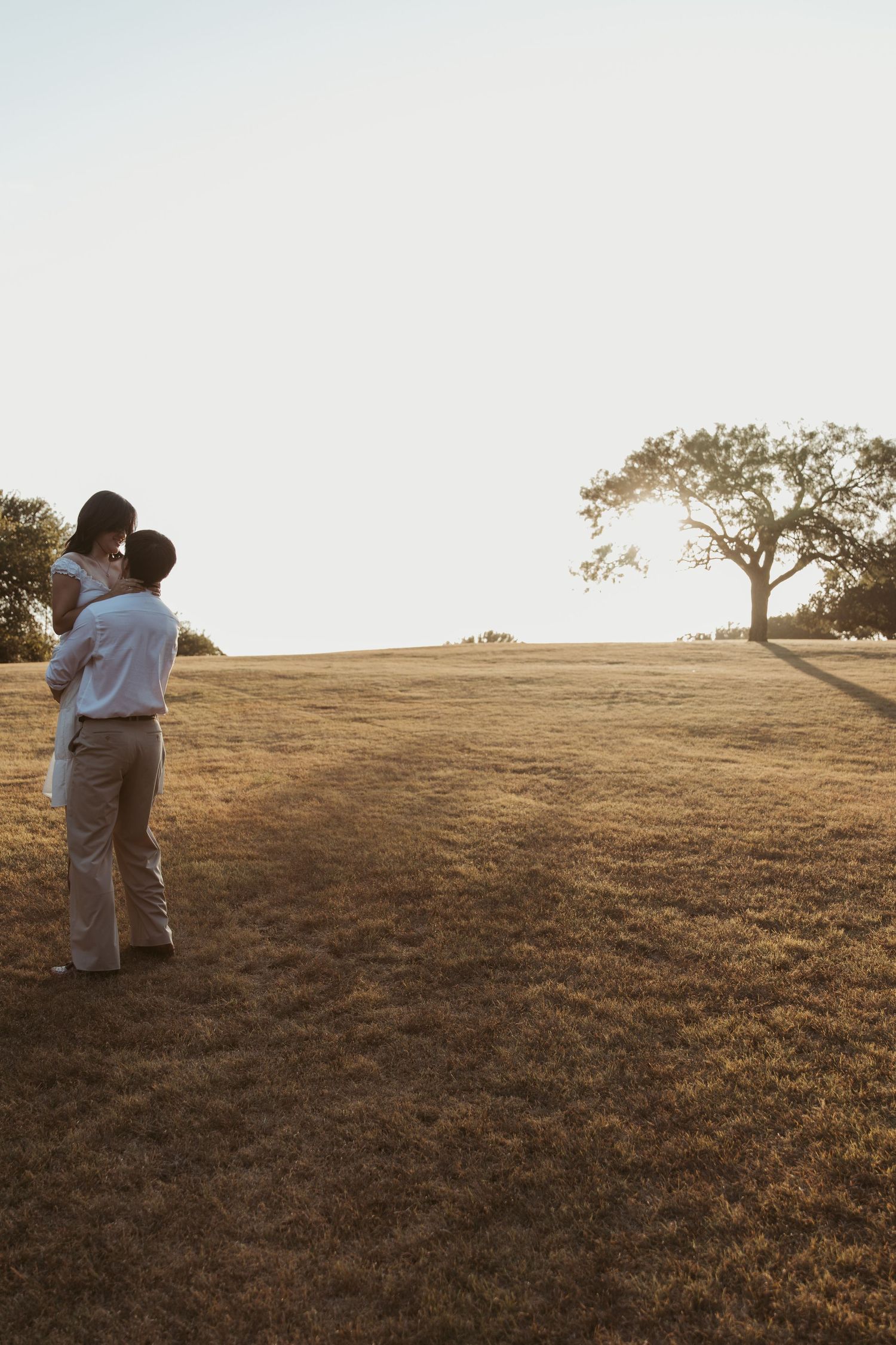 A couple shares an intimate moment at sunset on a grassy hilltop with golden light in the background.