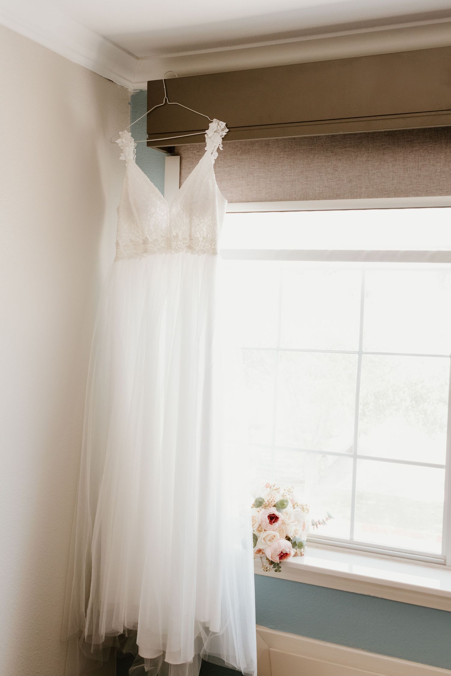 A white wedding dress hangs elegantly by a window with a delicate bouquet of pink and white flowers nearby.