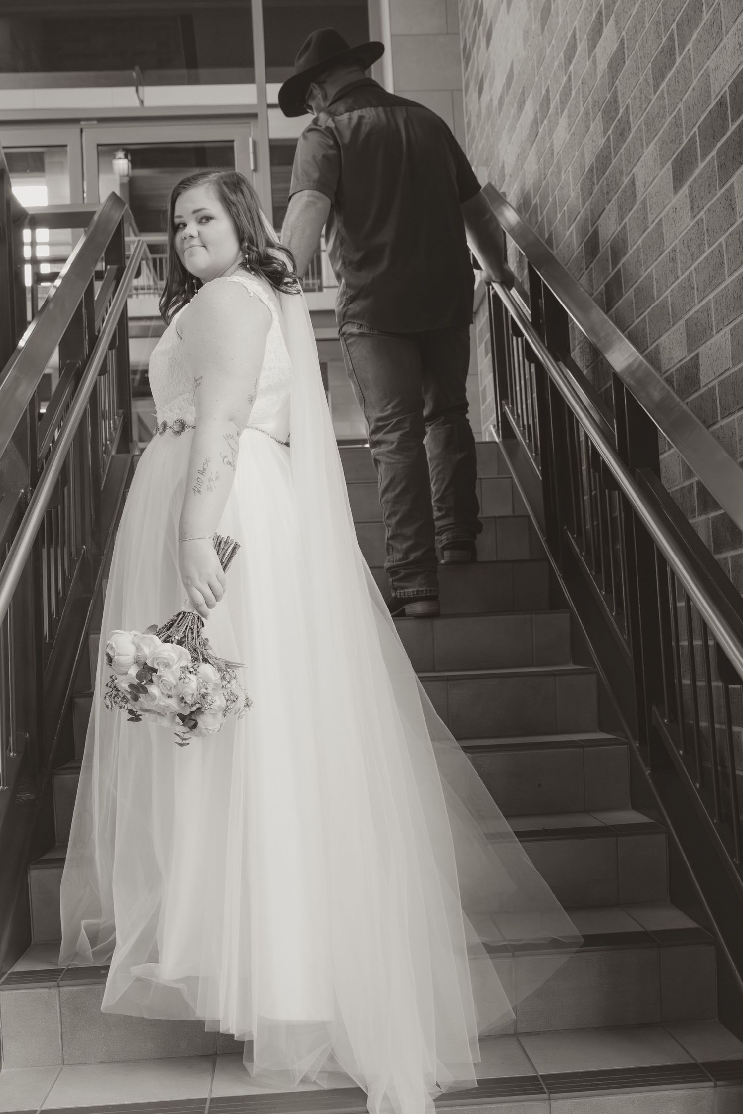 A bride in a flowing white dress poses on stairs while her partner stands ahead wearing a black hat.