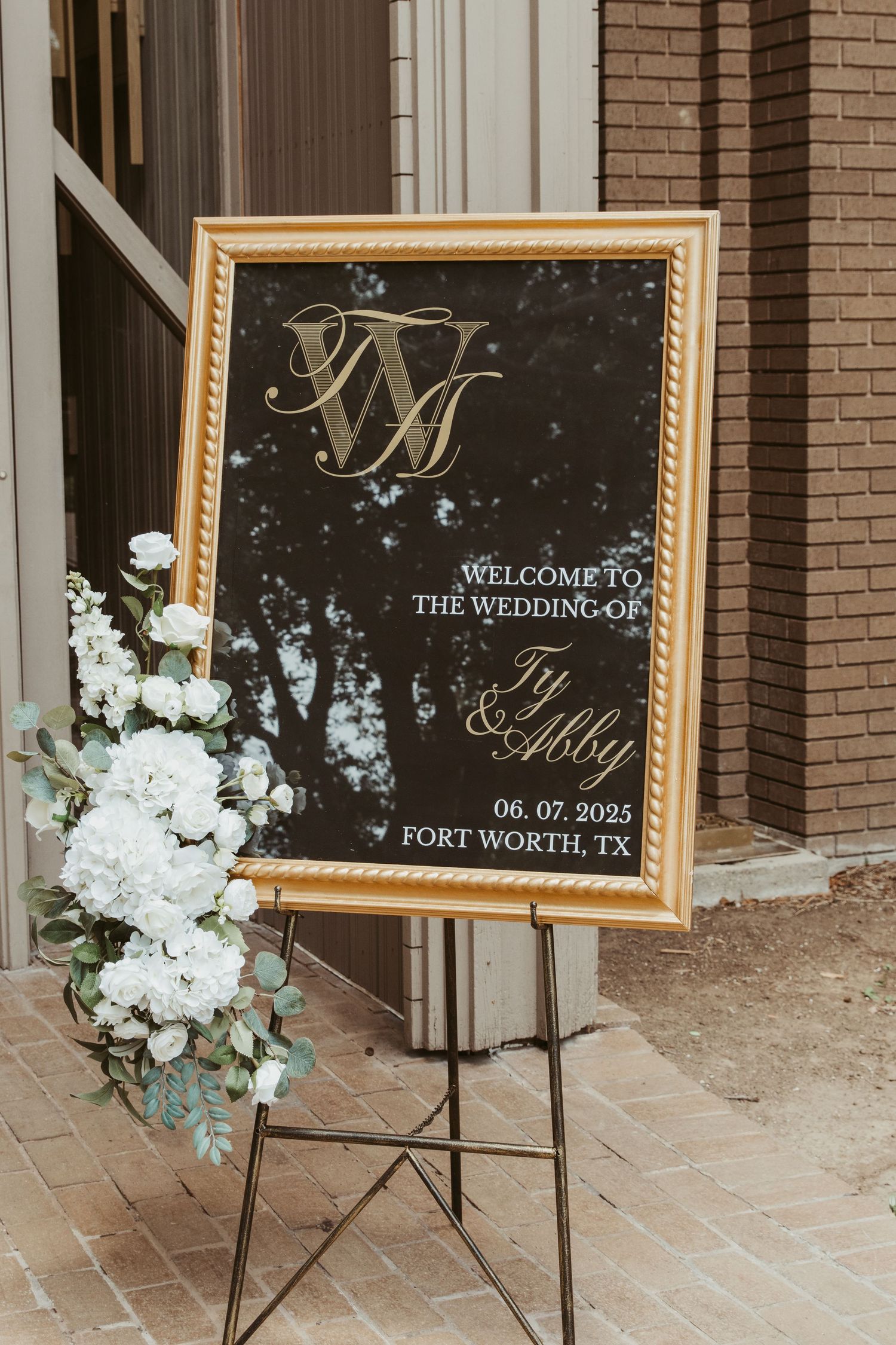 Elegant gold-framed wedding welcome sign with floral decorations displayed on an easel outside a brick venue in Fort Worth.