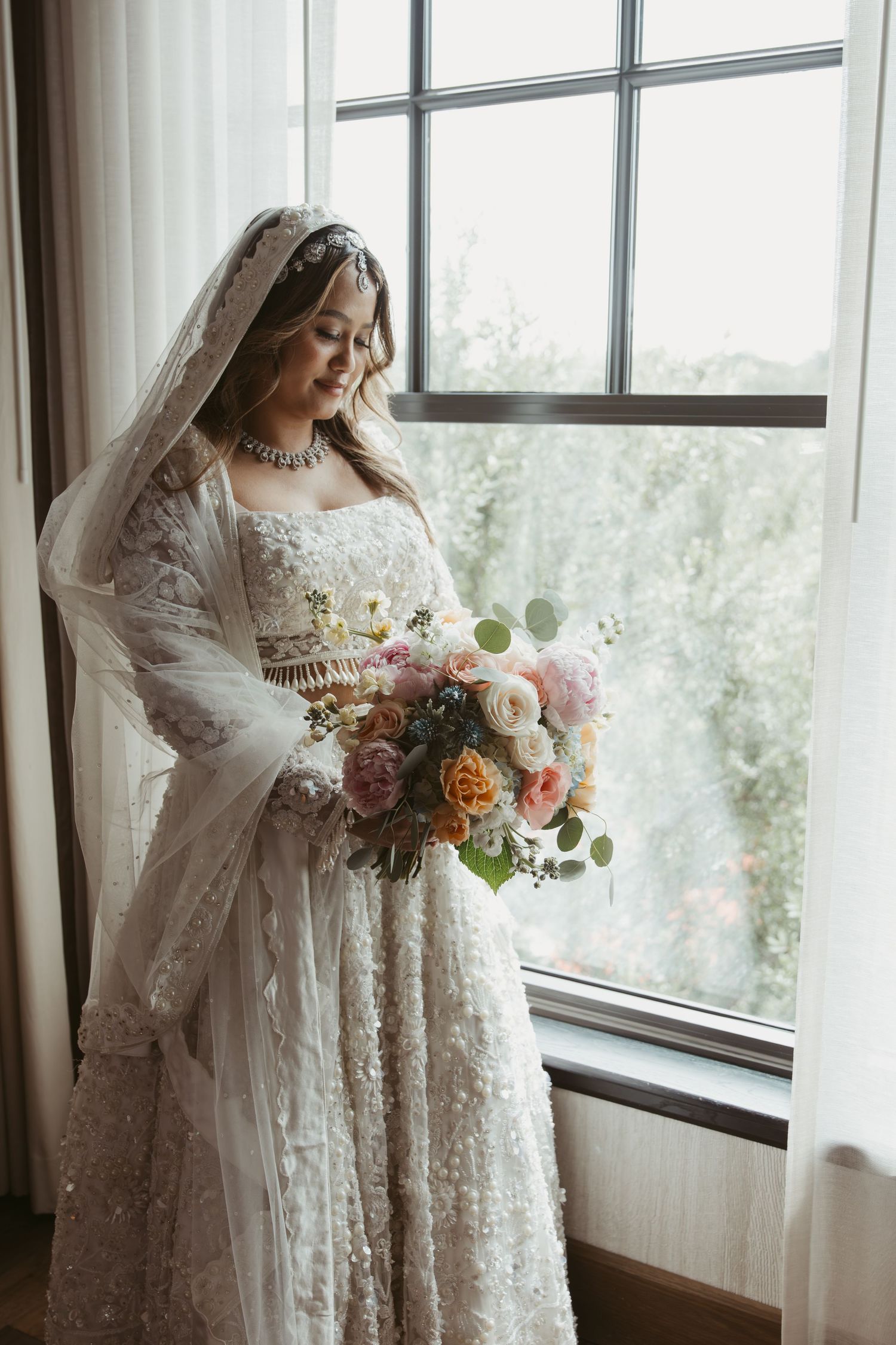A bride in a vintage lace wedding dress stands by a window holding a lush bouquet of pink and peach flowers.