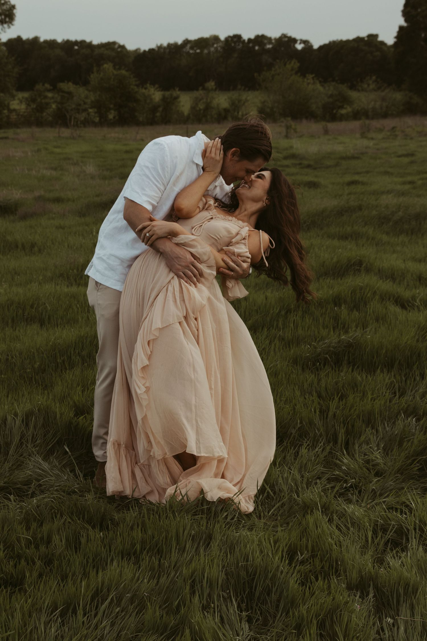 A romantic couple embraces in a dreamy meadow at dusk, with her wearing a flowing blush gown and him in white attire.