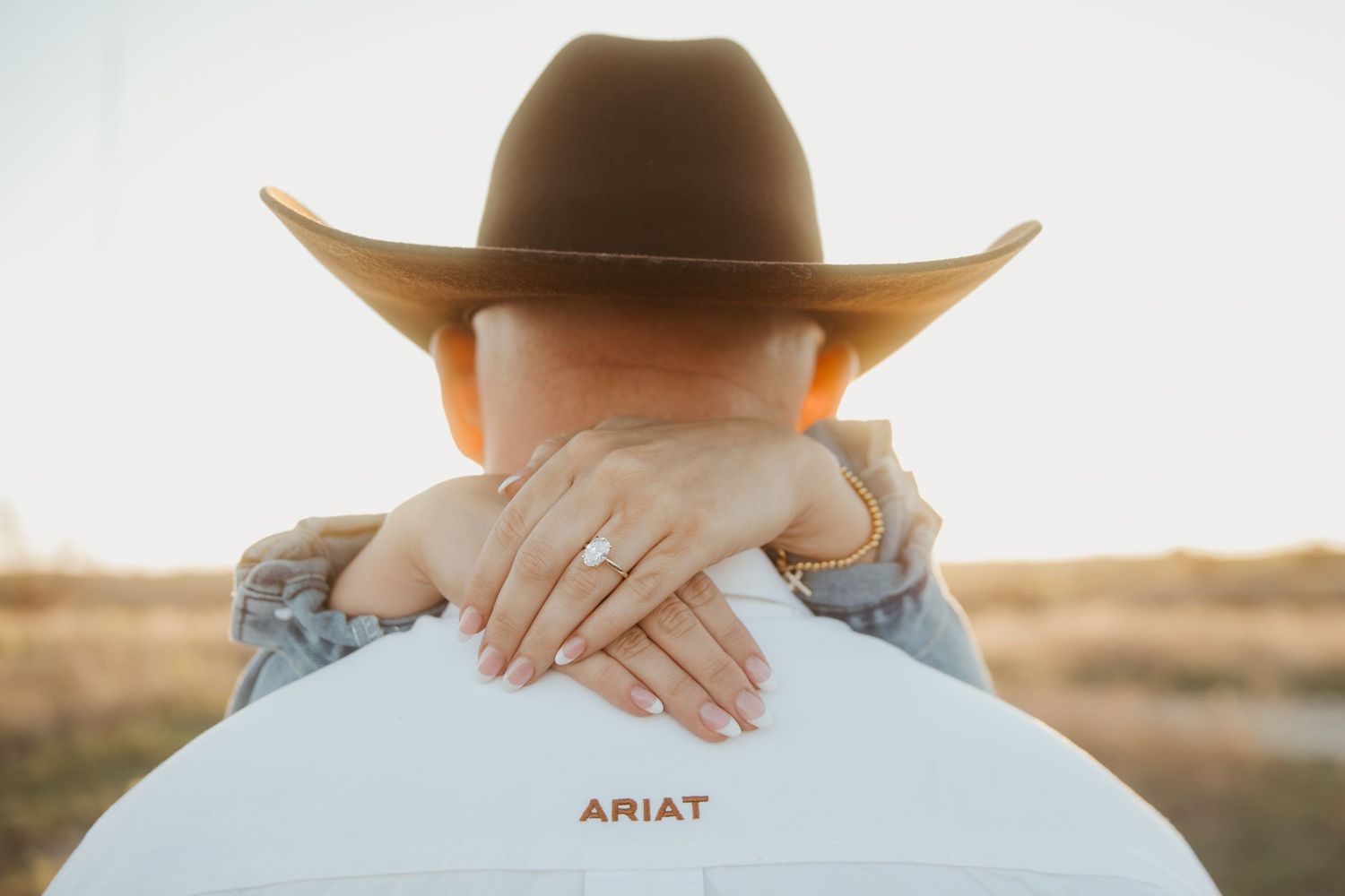 Woman in cowboy hat embracing partner wearing Ariat shirt, showing off engagement ring at golden hour.
