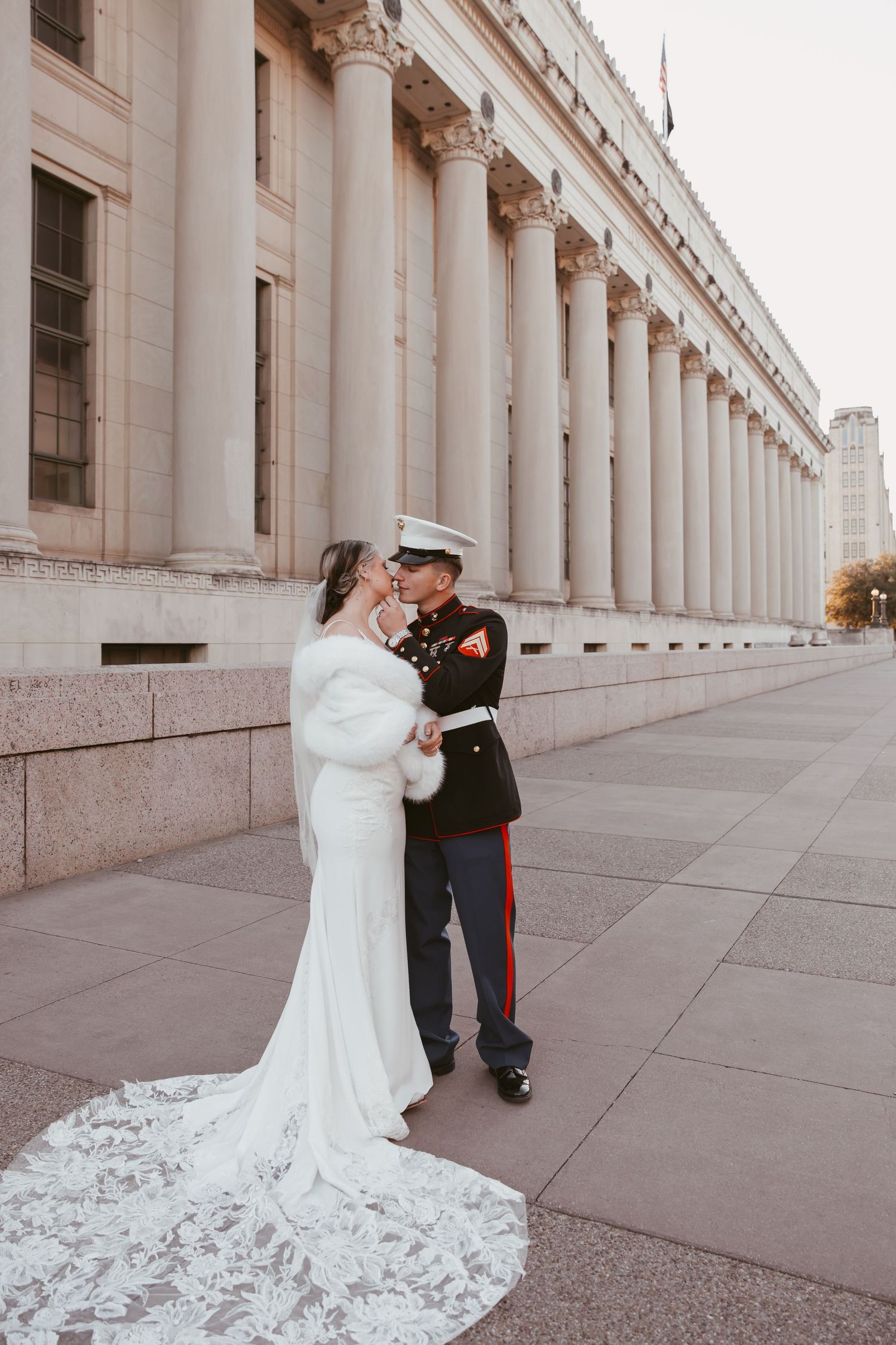 A couple in wedding attire embrace outside a grand neoclassical building with tall white columns.
