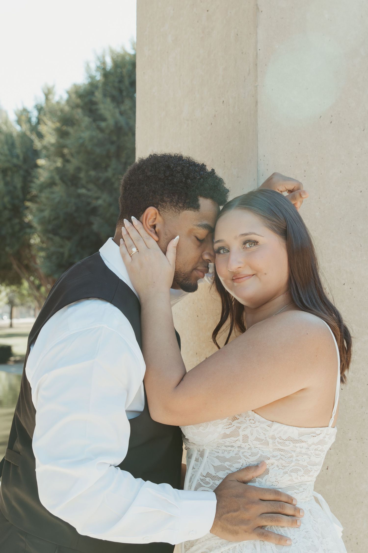 A couple embraces tenderly against a stone pillar with trees in the background. Wedding Photographer DFW