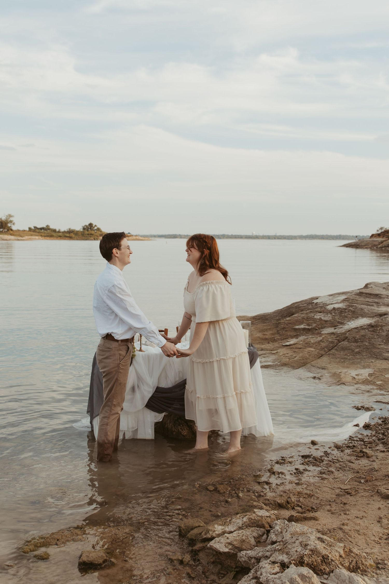 A romantic proposal scene unfolds on a rocky beach shoreline at sunset with two people in light-colored clothing.