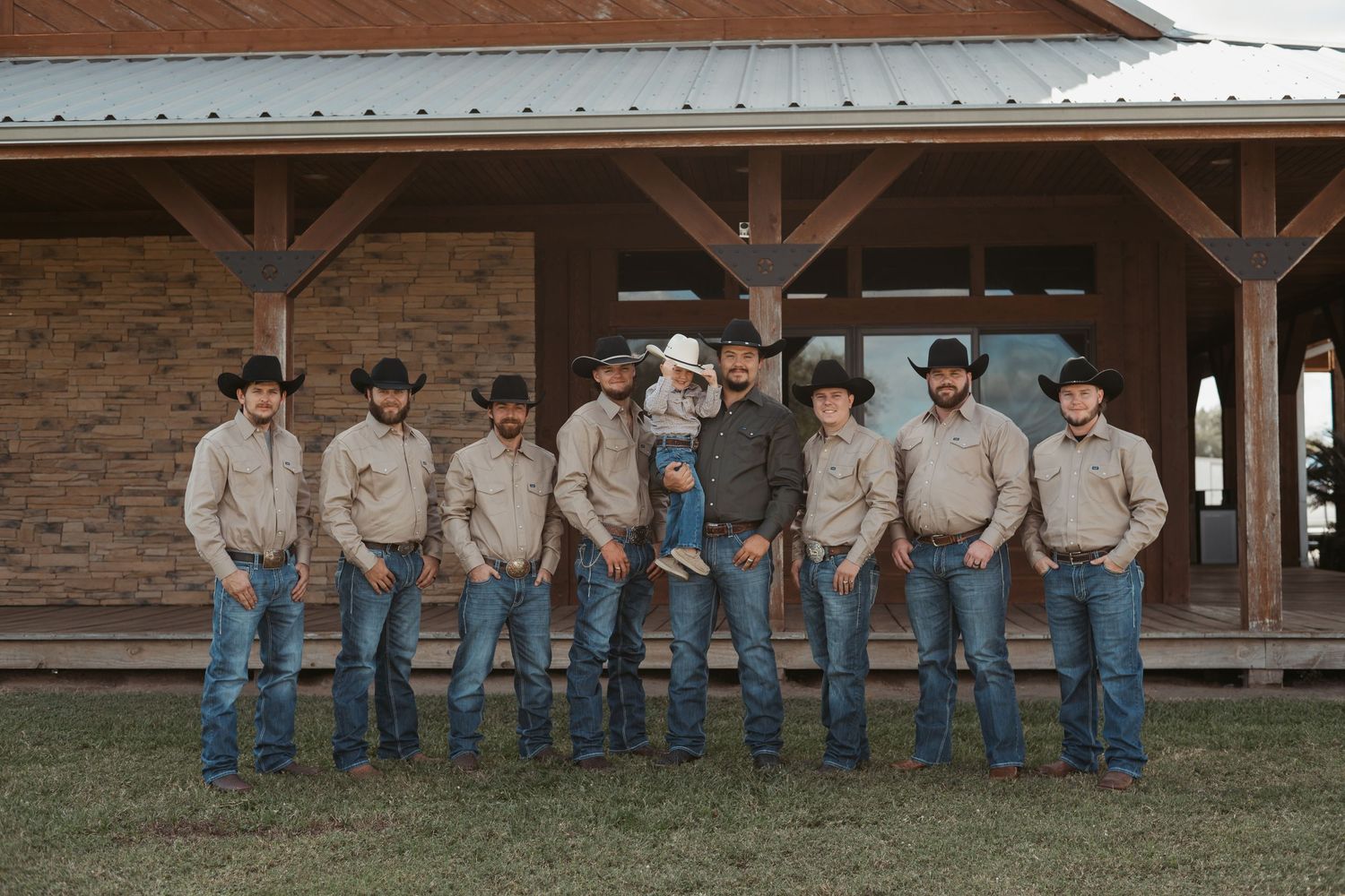 Group wearing matching beige shirts and jeans pose casually along rustic wooden porch railing at sunset. Wedding photographer DFW
