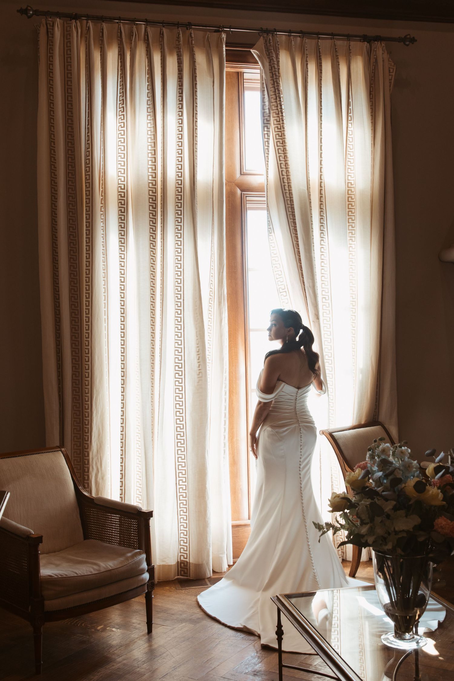 A silhouetted figure in a white dress stands near sunlit curtained windows in a serene interior setting.