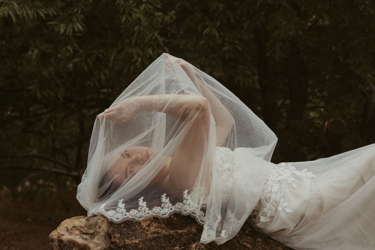A flowing white wedding dress train draped across dark forest ground creates an ethereal woodland scene.