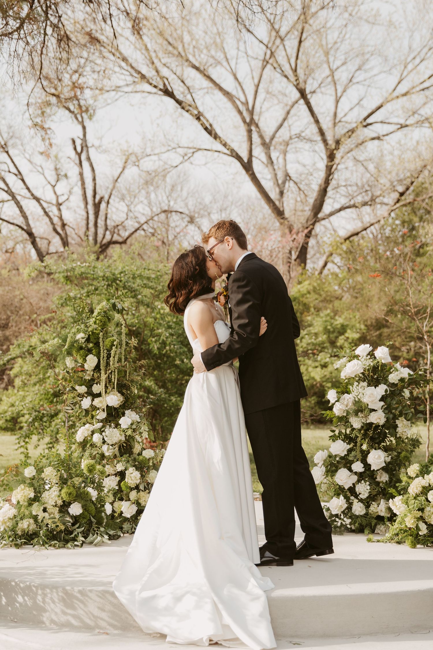 Bride and groom share first kiss at outdoor wedding altar surrounded by white floral arrangements.