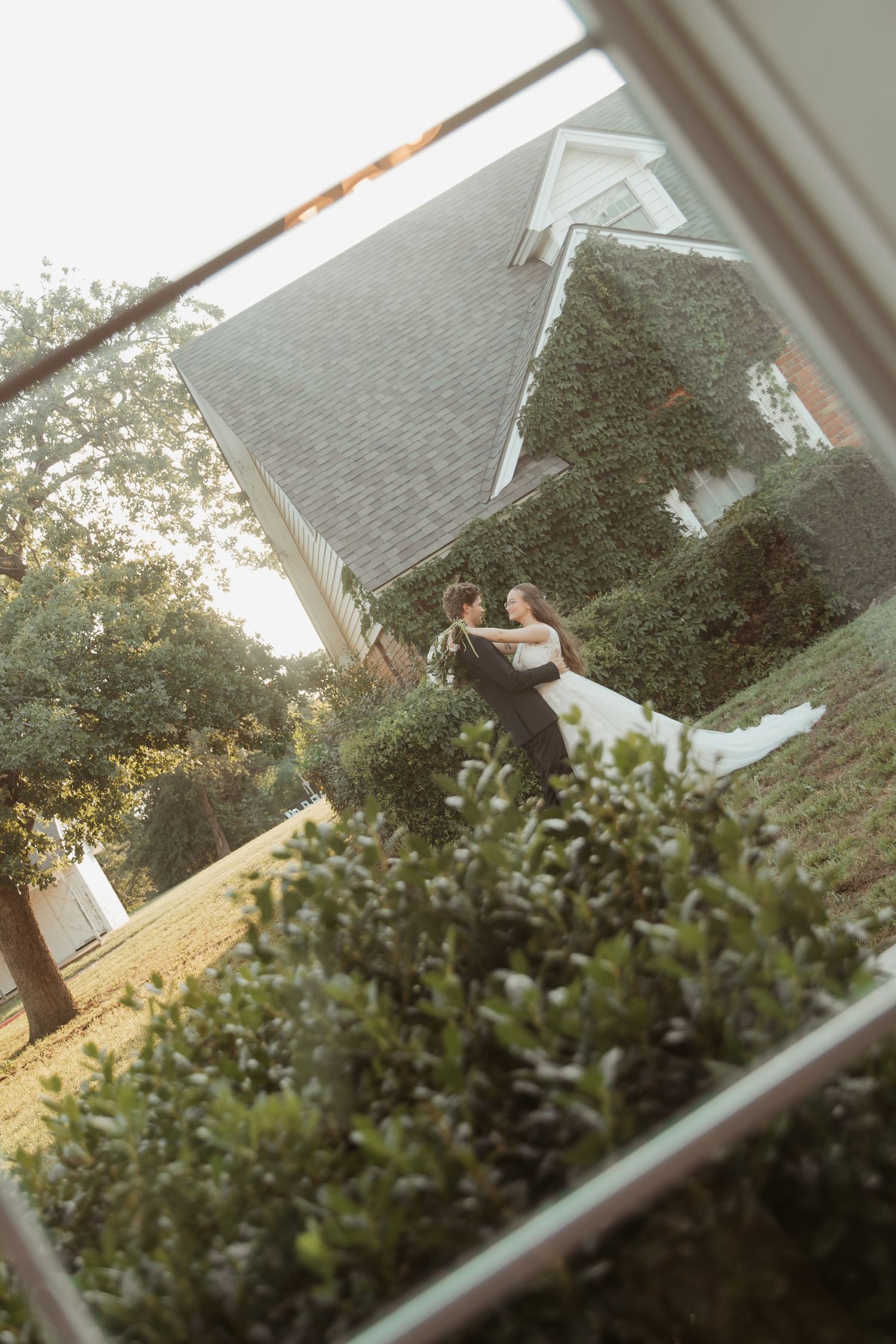 A bride and groom embrace in a lush garden outside an ivy-covered brick building, captured through a window frame.