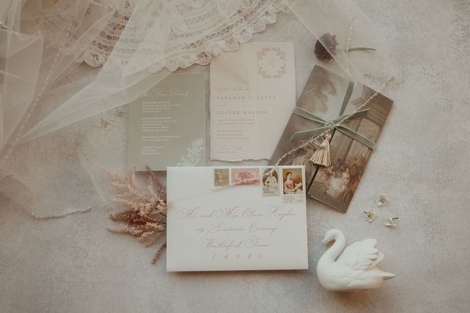 Elegant wedding invitation suite with envelope, stamps, lace veil, dried florals, and a white swan figurine on a neutral background.
