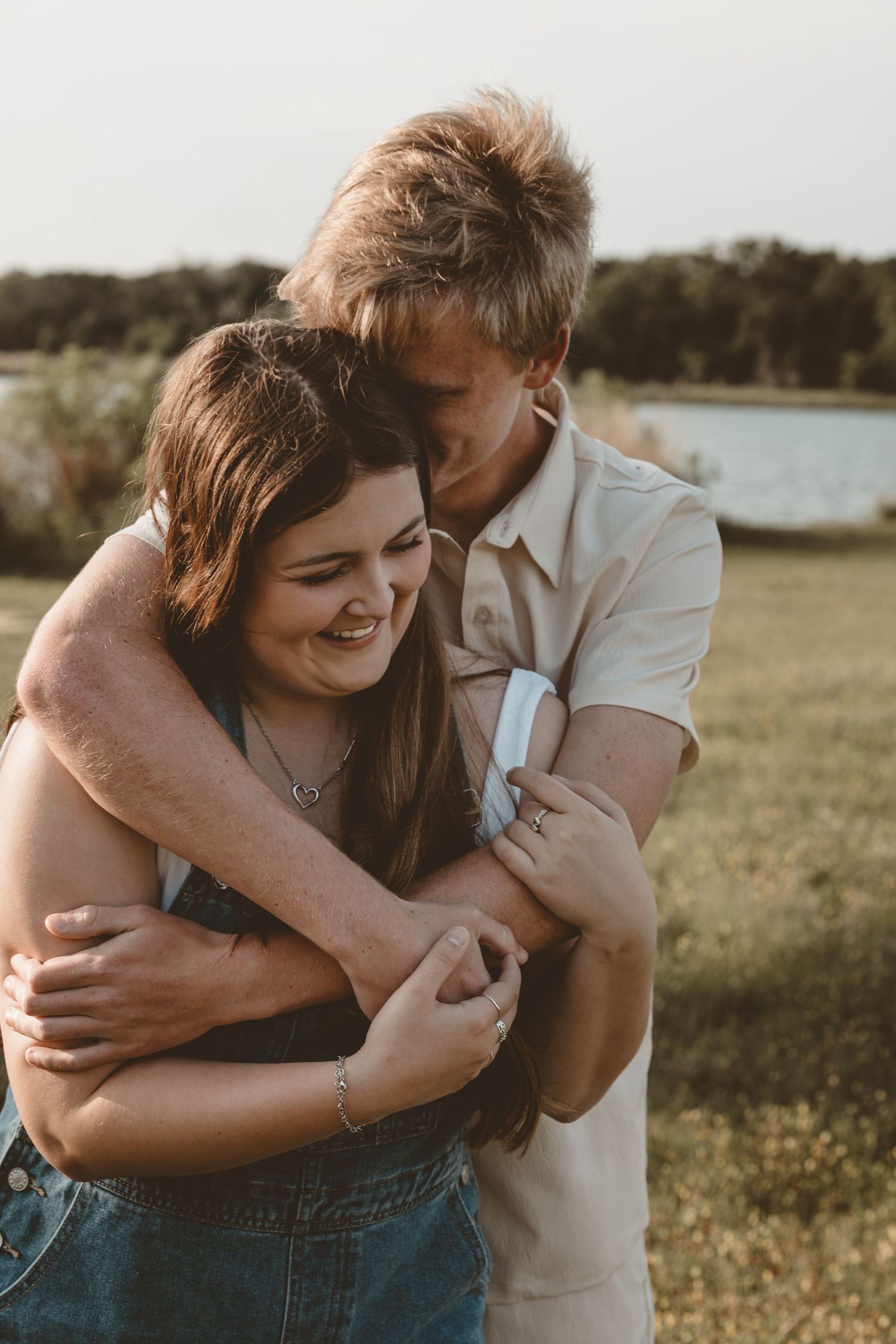Affectionate couple embracing in a sun-lit field near water during golden hour.