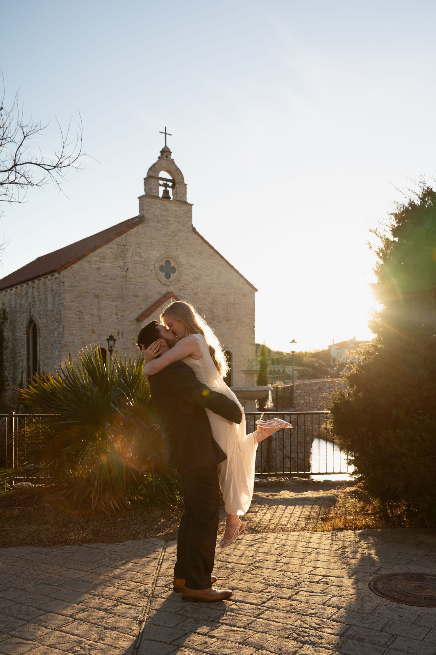 A romantic sunset silhouette of a couple embracing outside a rustic stone church with a bell tower and cross.