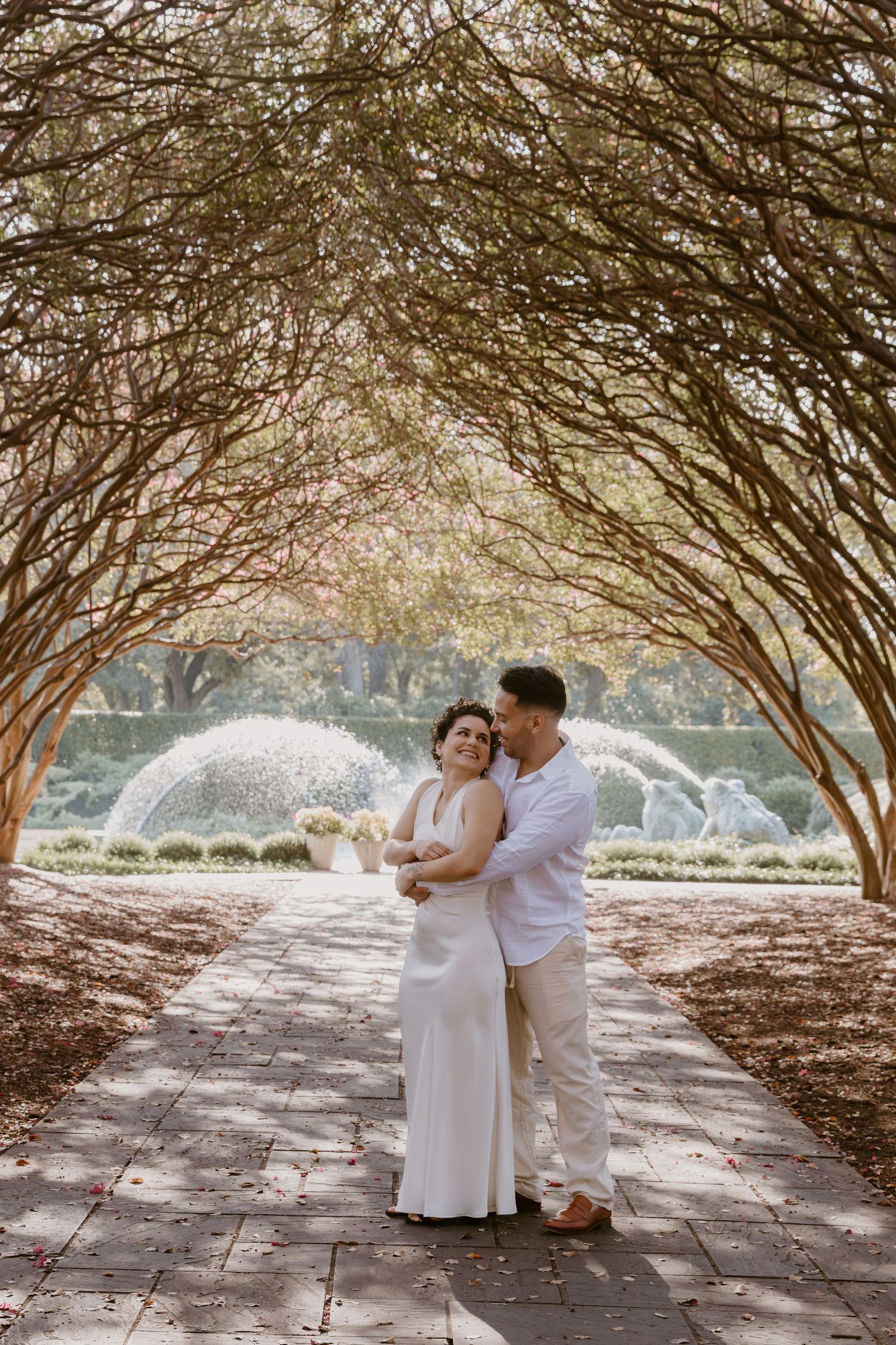 A romantic engagement photo session under a canopy of trees with natural sunlight filtering through the branches.