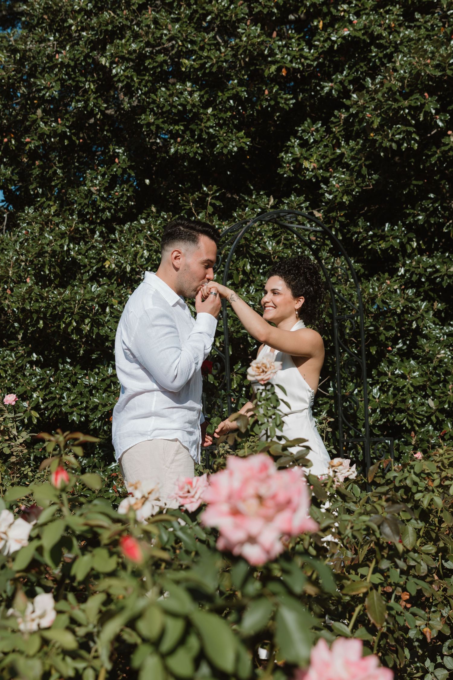 A couple shares a romantic moment among blooming pink roses in a garden setting.