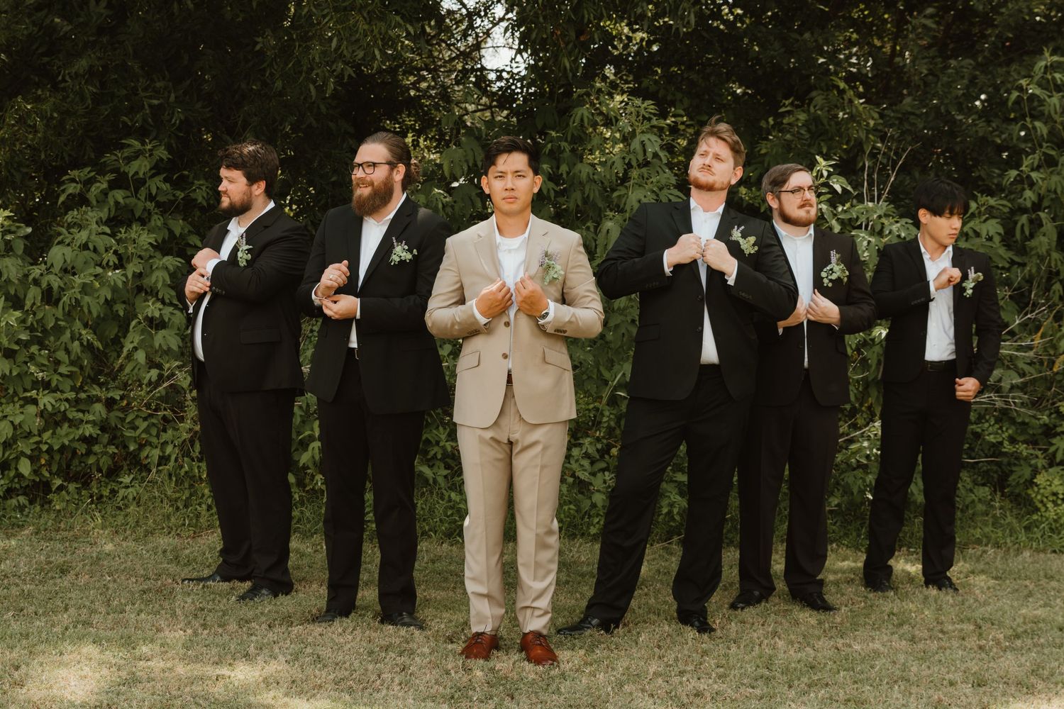 Wedding party members in formal attire standing in a line outdoors against greenery.