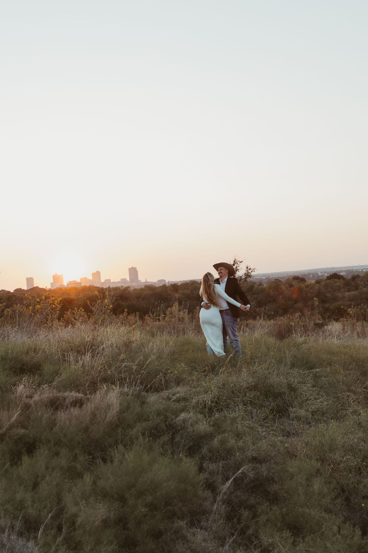 A couple dances together on a hilltop at sunset with a city skyline visible in the background.
