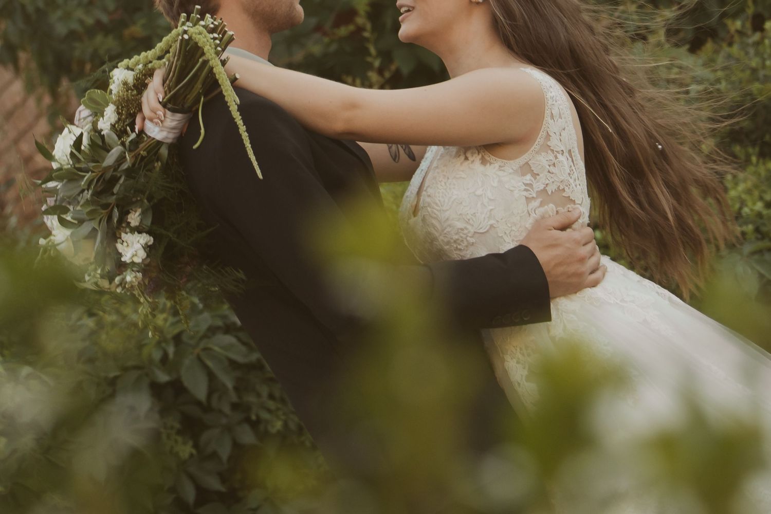 A bride in a lace gown embraces a groom in a dark suit outdoors, holding a lush green floral bouquet.