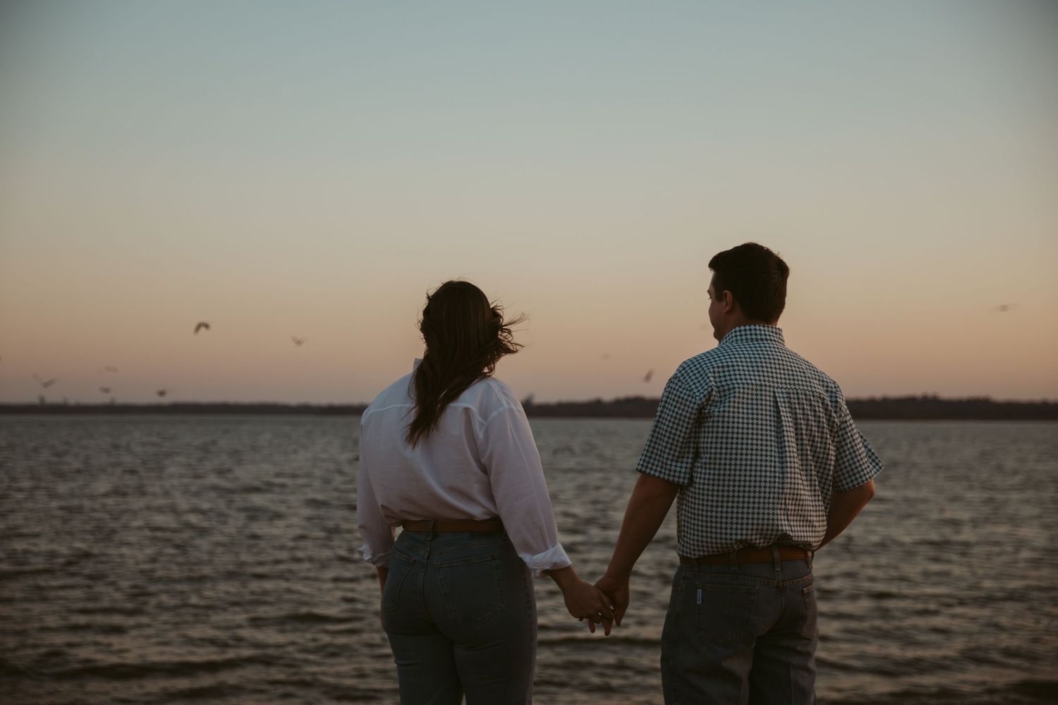 Couple holding hands facing the ocean at a colorful dusk sunset by the water.